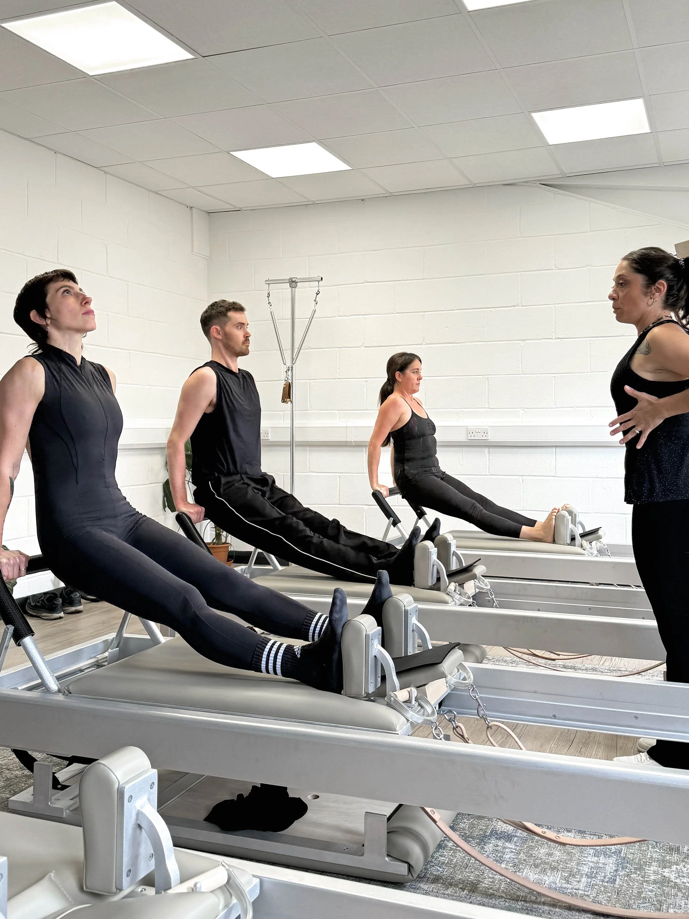 Four people participating in a Pilates class on reformer machines, with an teacher guiding them in a bright room with white walls and ceiling lights.