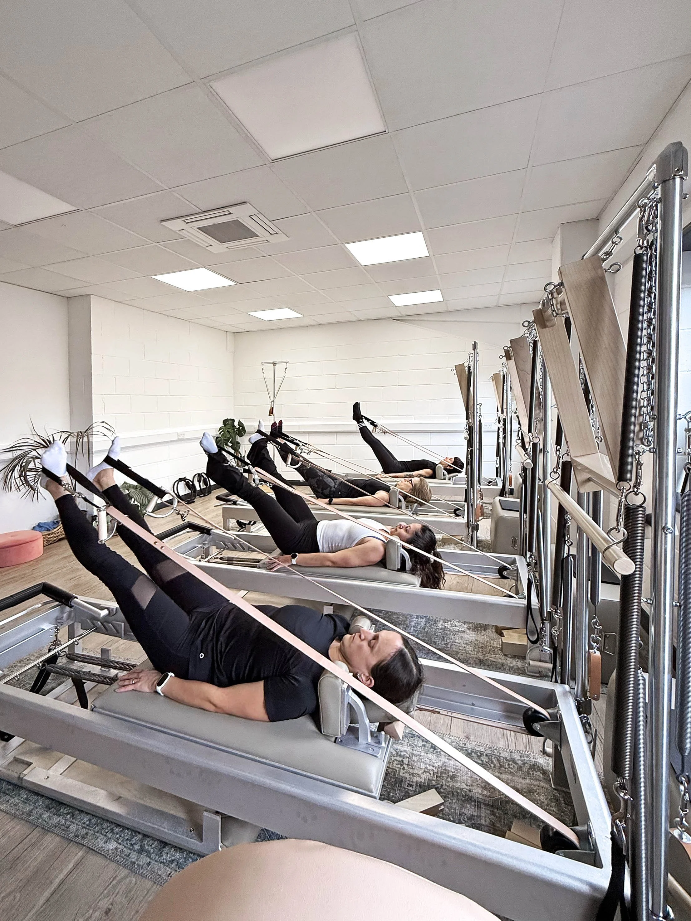 Four women exercising on Pilates reformer machines in a fitness studio, lying on their backs with legs raised and connected to resistance bands.