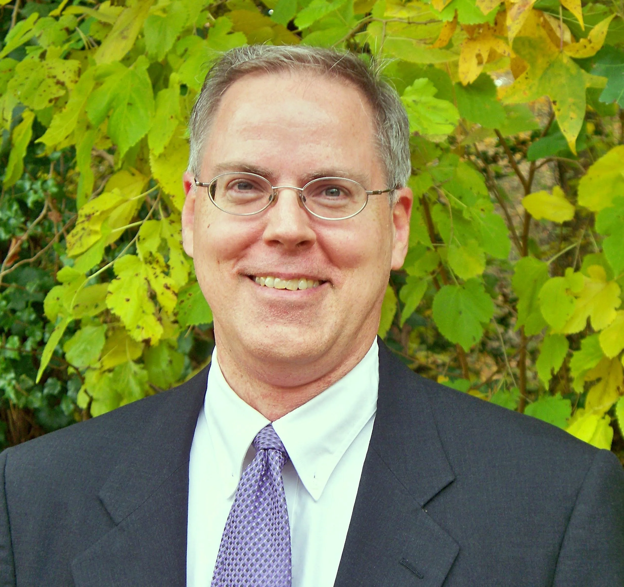 A man smiling, wearing glasses and a suit with a purple tie, standing outdoors in front of green foliage.