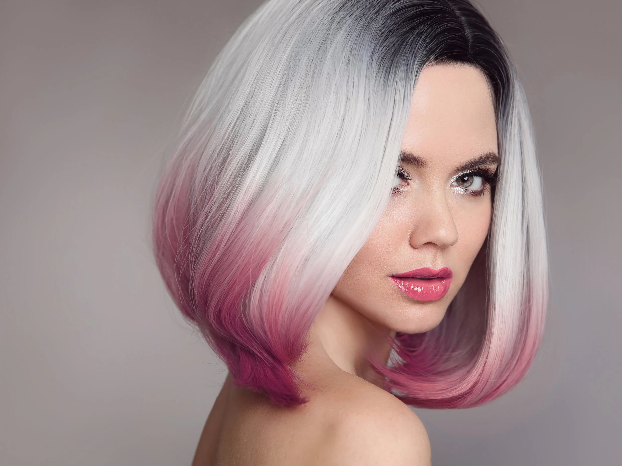 A woman with short hair styled in ombre colors, starting from black at the roots, fading into white, and ending with pink at the tips. She has makeup with glossy pink lipstick and subtle eye makeup, and looks over her shoulder.