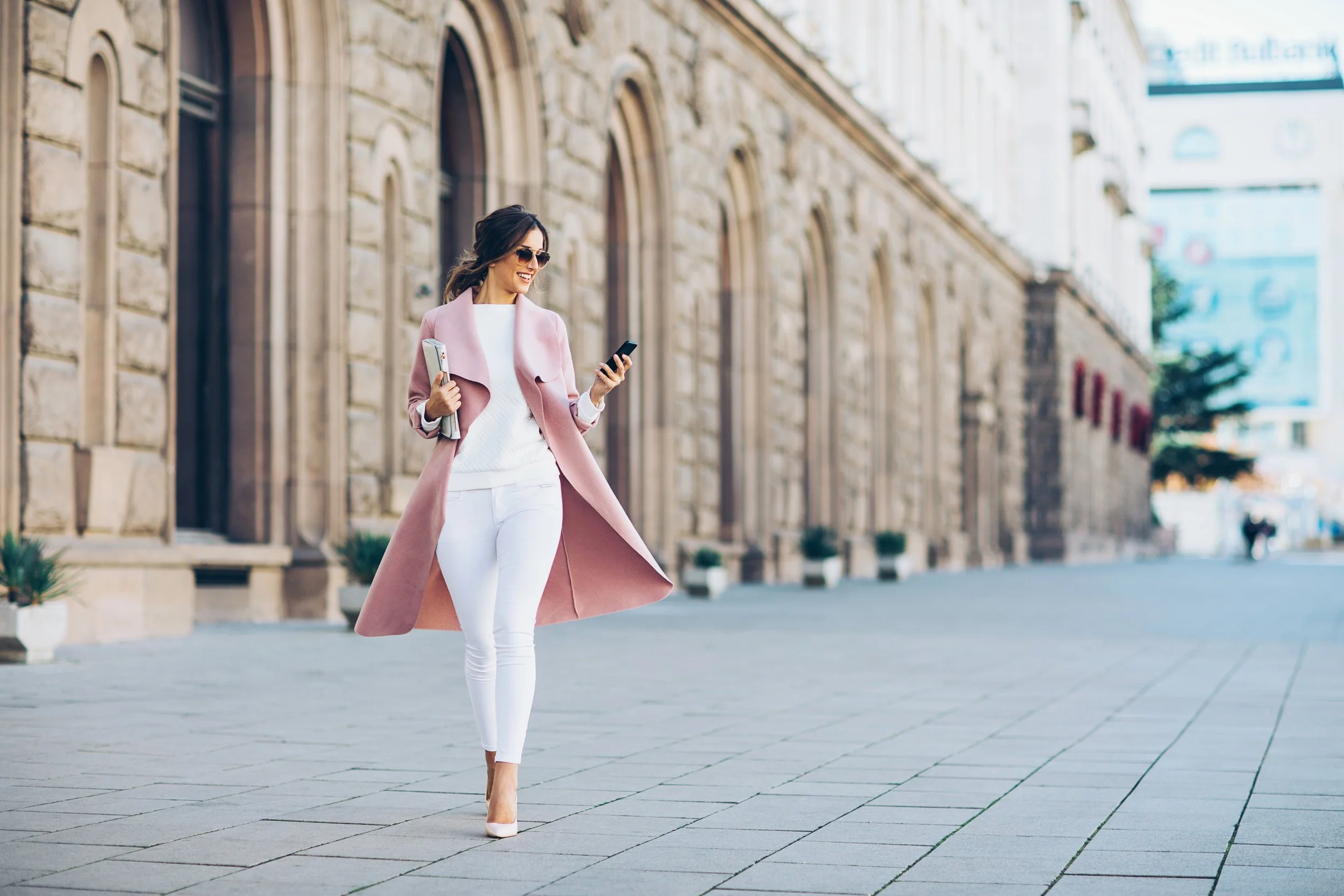 A woman walking on a city street, dressed stylishly in a pink coat, white pants, and heels, holding a smartphone and a magazine, smiling as she looks at her phone.