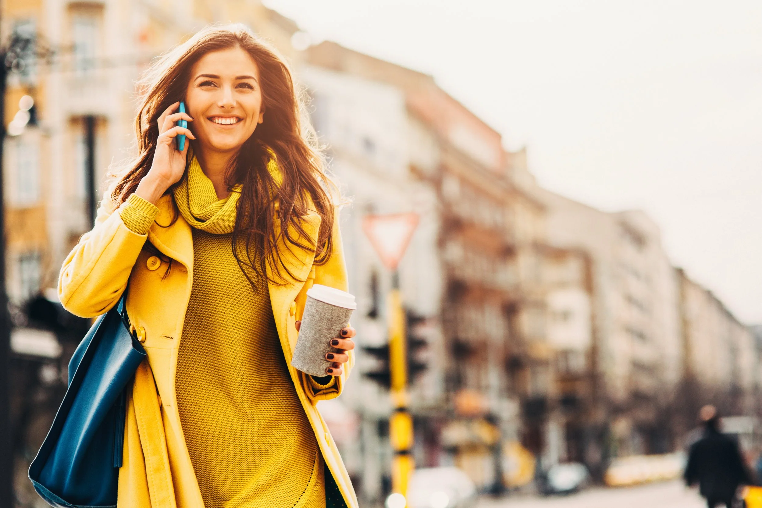 A woman with long brown hair smiling and talking on a cell phone while walking down a city street, holding a coffee cup, dressed in a yellow coat and sweater, with a blue tote bag.