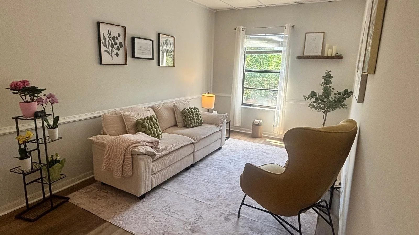 Living room with beige sofa decorated with two green pillows, a beige armchair, a side table with a lamp, and potted plants. There are framed pictures on the wall and a window with white curtains, allowing natural sunlight.