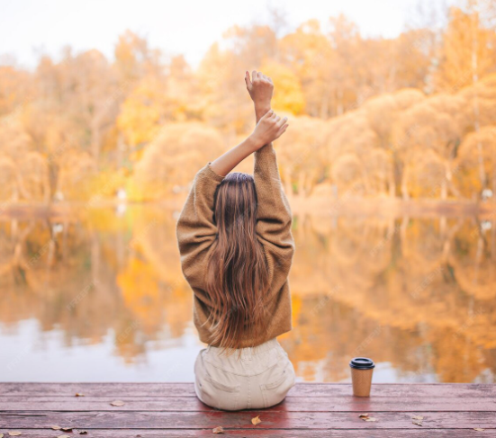 Mujer sentada en un muelle, mirando un lago con árboles en tonos otoñales, sosteniendo sus brazos en señal de relajación, con una taza de café a su lado.