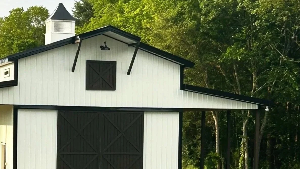 White barn with black trim and large sliding doors, set against a backdrop of green trees.
