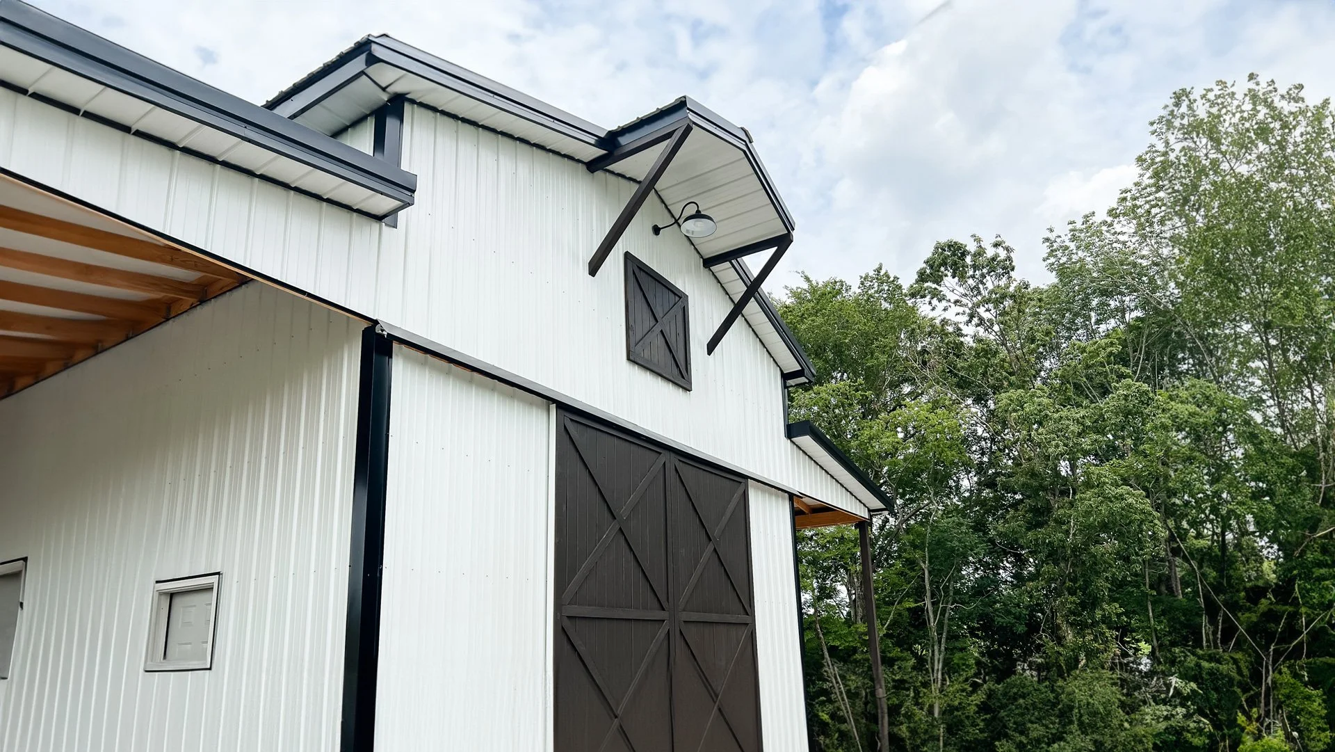 A modern barn with white corrugated metal siding, black accents, and large sliding doors, set against a background of green trees and a cloudy sky.