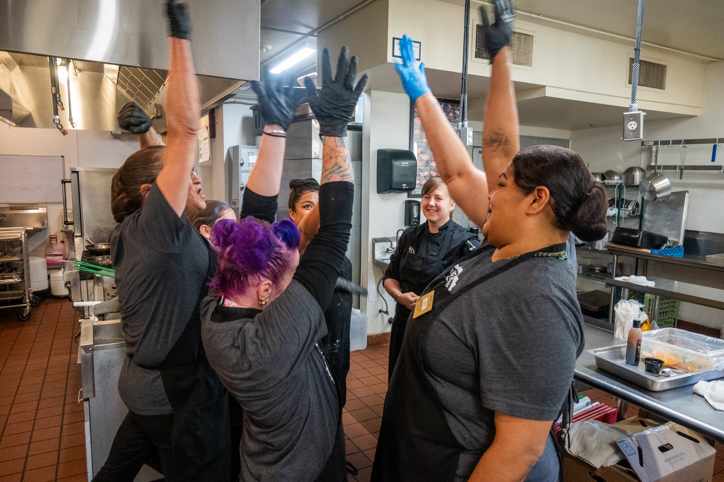 Group of women wearing aprons standing in a commercial kitchen raising their hands in the air in celebration