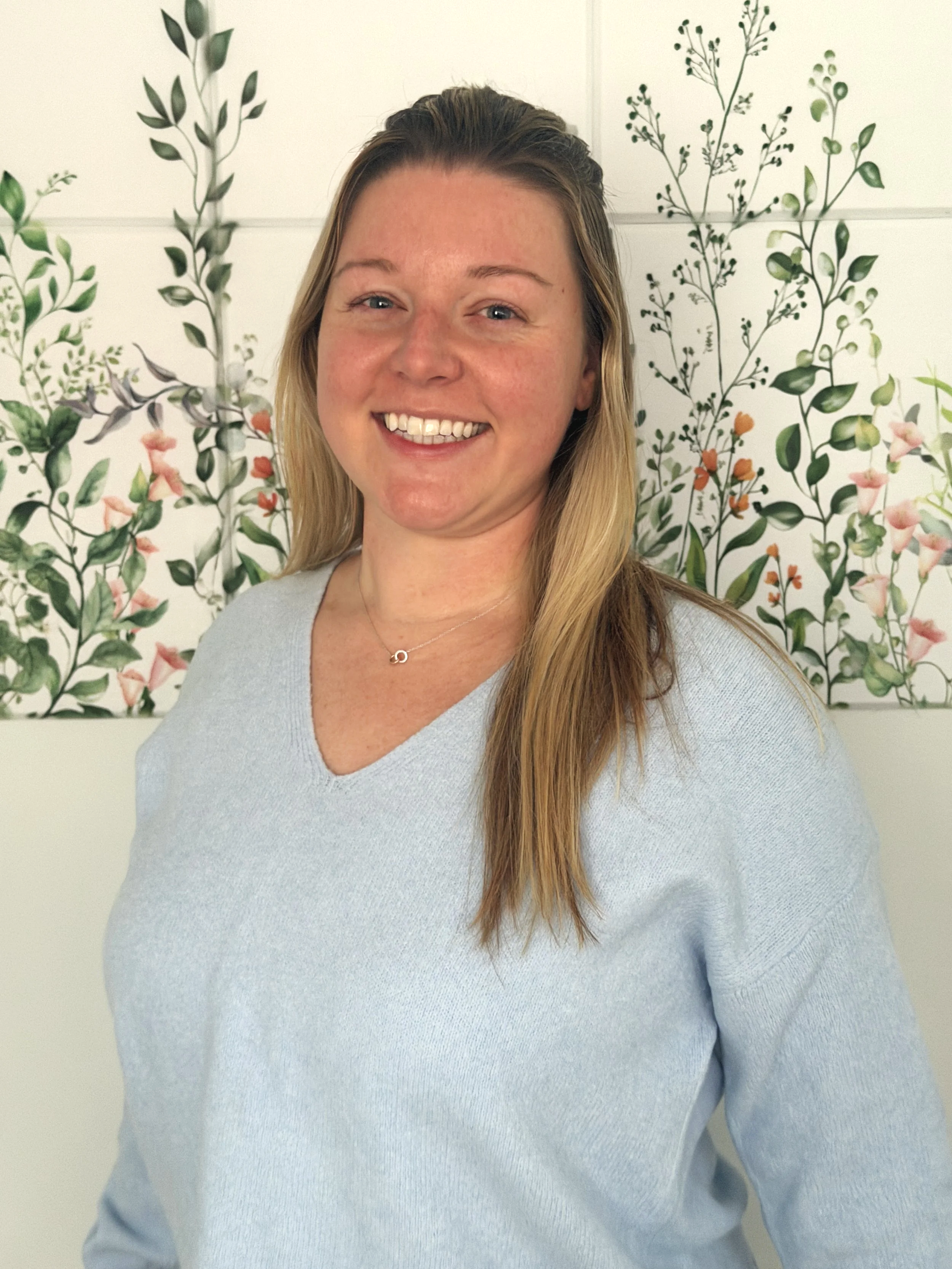 A woman with long blond hair smiling in front of a wall with floral artwork.