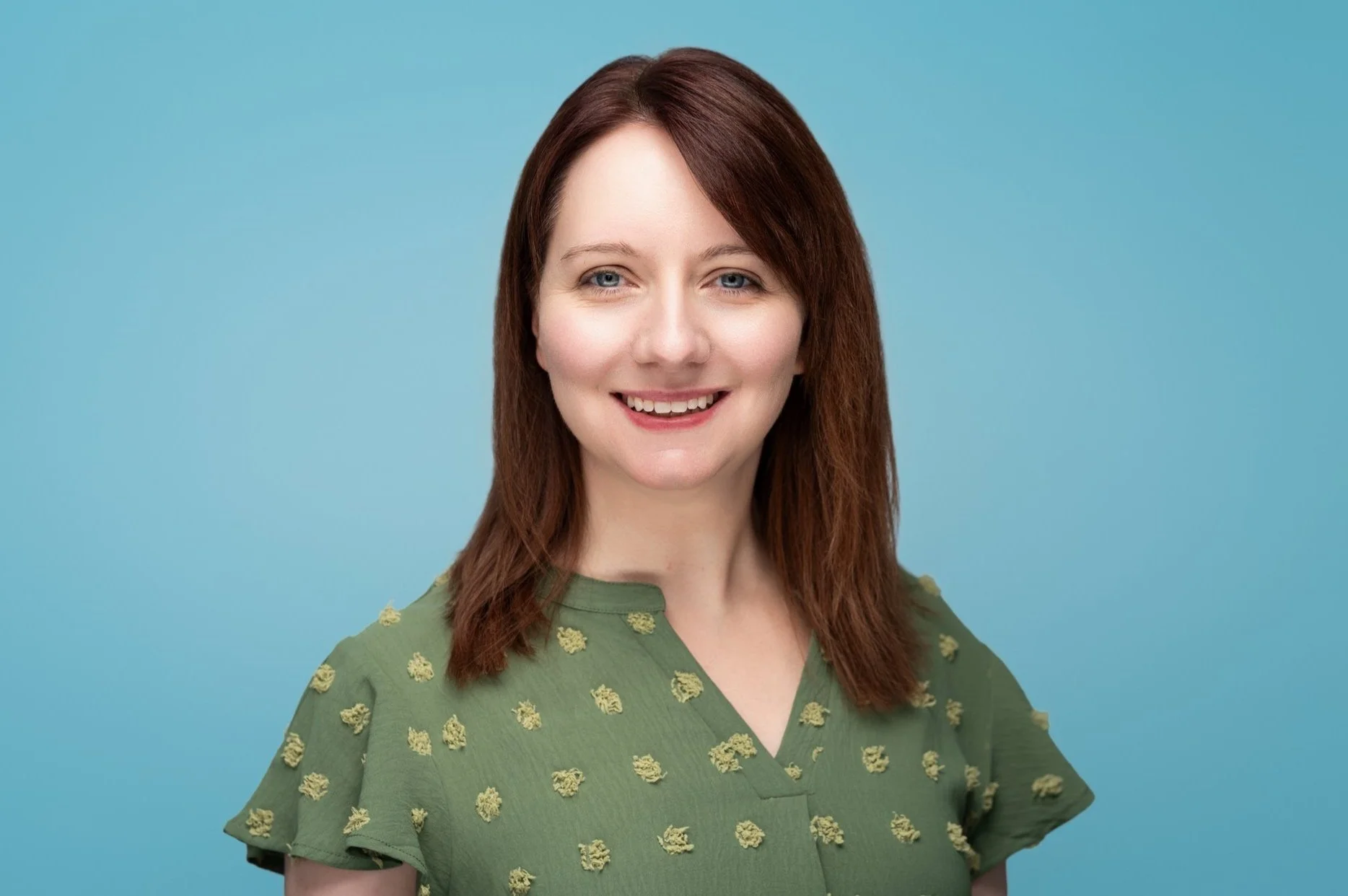 Portrait of a woman with shoulder-length brown hair, blue eyes, and fair skin smiling, wearing a green top with small yellow floral patterns, against a plain blue background.