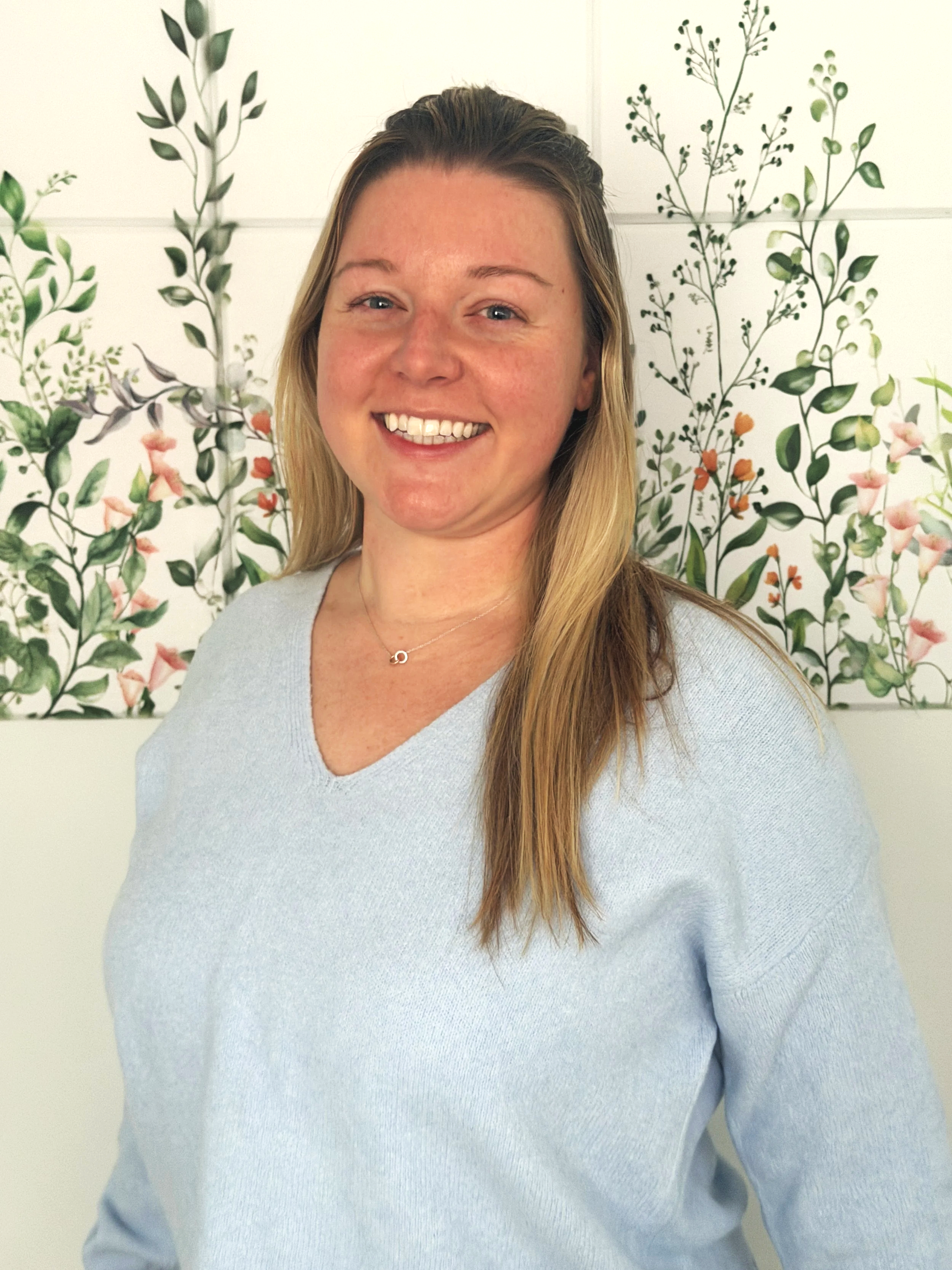 A smiling woman with long blonde hair wearing a light blue sweater and a necklace, standing in front of a wall decorated with floral artwork.