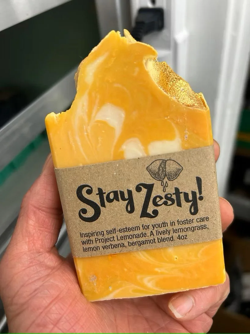 Person holding "Squeeze the Day" soap bar in front of shelving with various soap products.