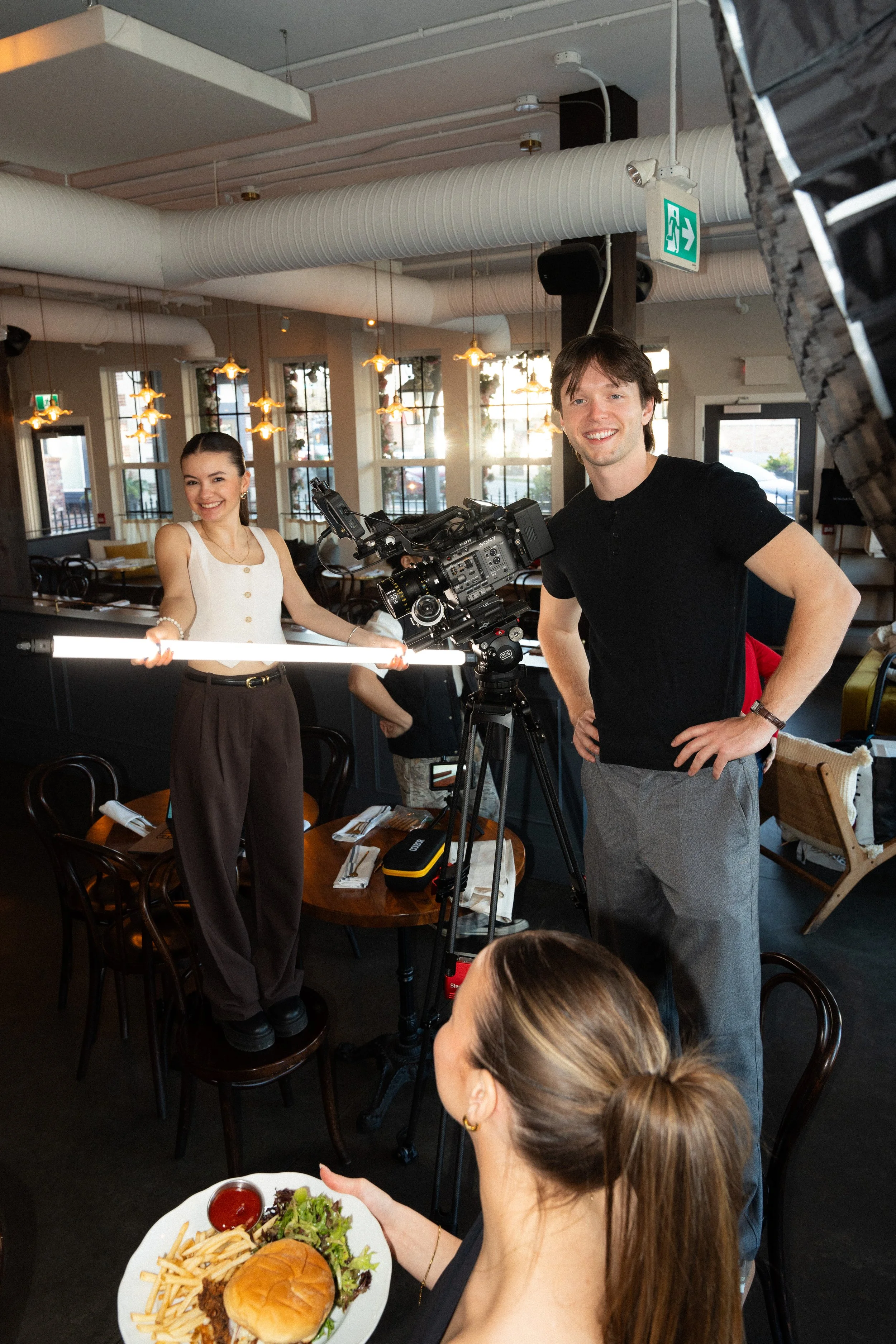 A woman with brown hair tied back holding a white plate with a burger, fries, salad, and condiments while a man with dark hair and a black shirt stands behind a professional camera set up on a tripod in a restaurant or cafe with natural light and warm hanging lights.