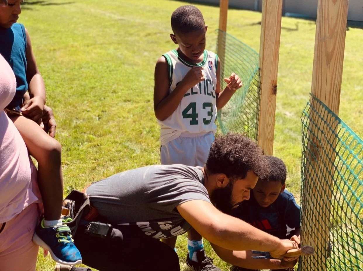 Group of people building a wooden fence with green mesh in a grassy area, including a child in a jersey and a man using tools.