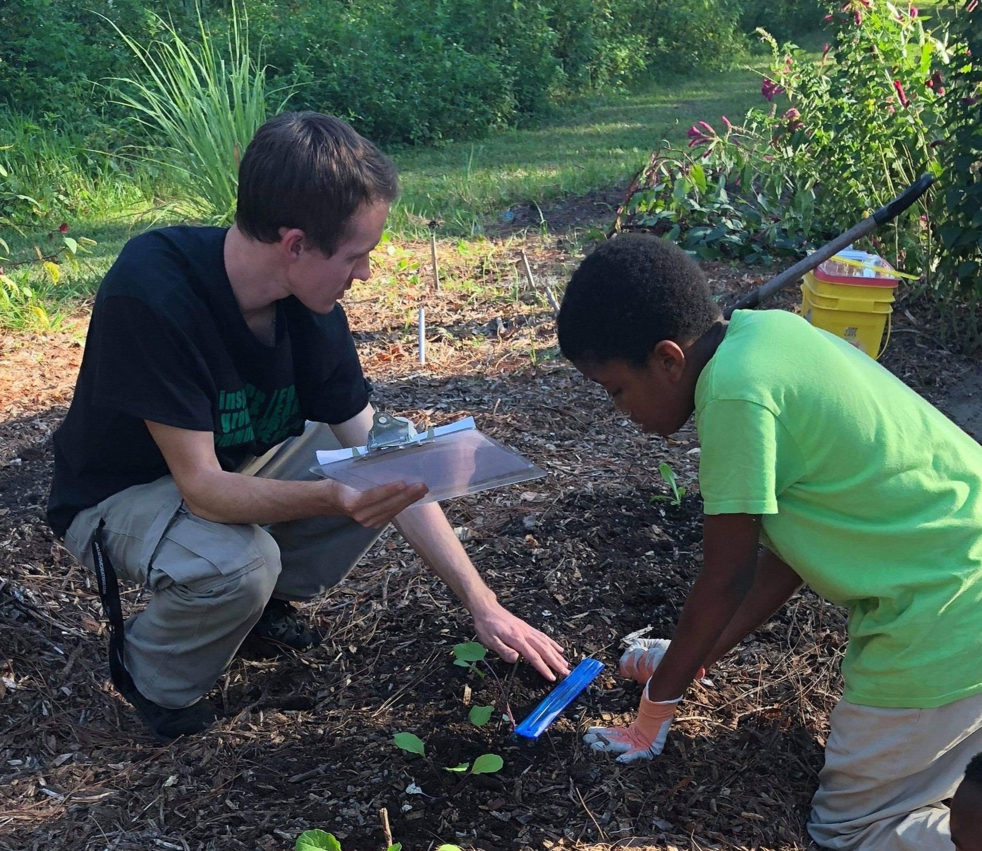 Two people gardening in an outdoor setting, one holding a clipboard and the other tending to the plants. They are surrounded by greenery and appear to be working together on a gardening task.