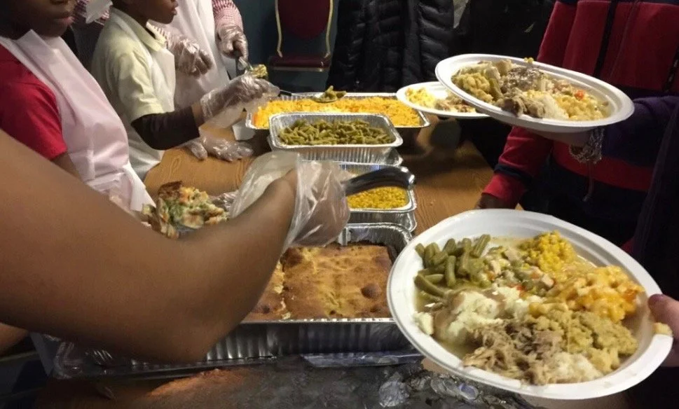 People serving food from buffet trays onto plates, including green beans, corn, cornbread, and meat dishes. Individuals wearing gloves, suggesting a meal service or community event.