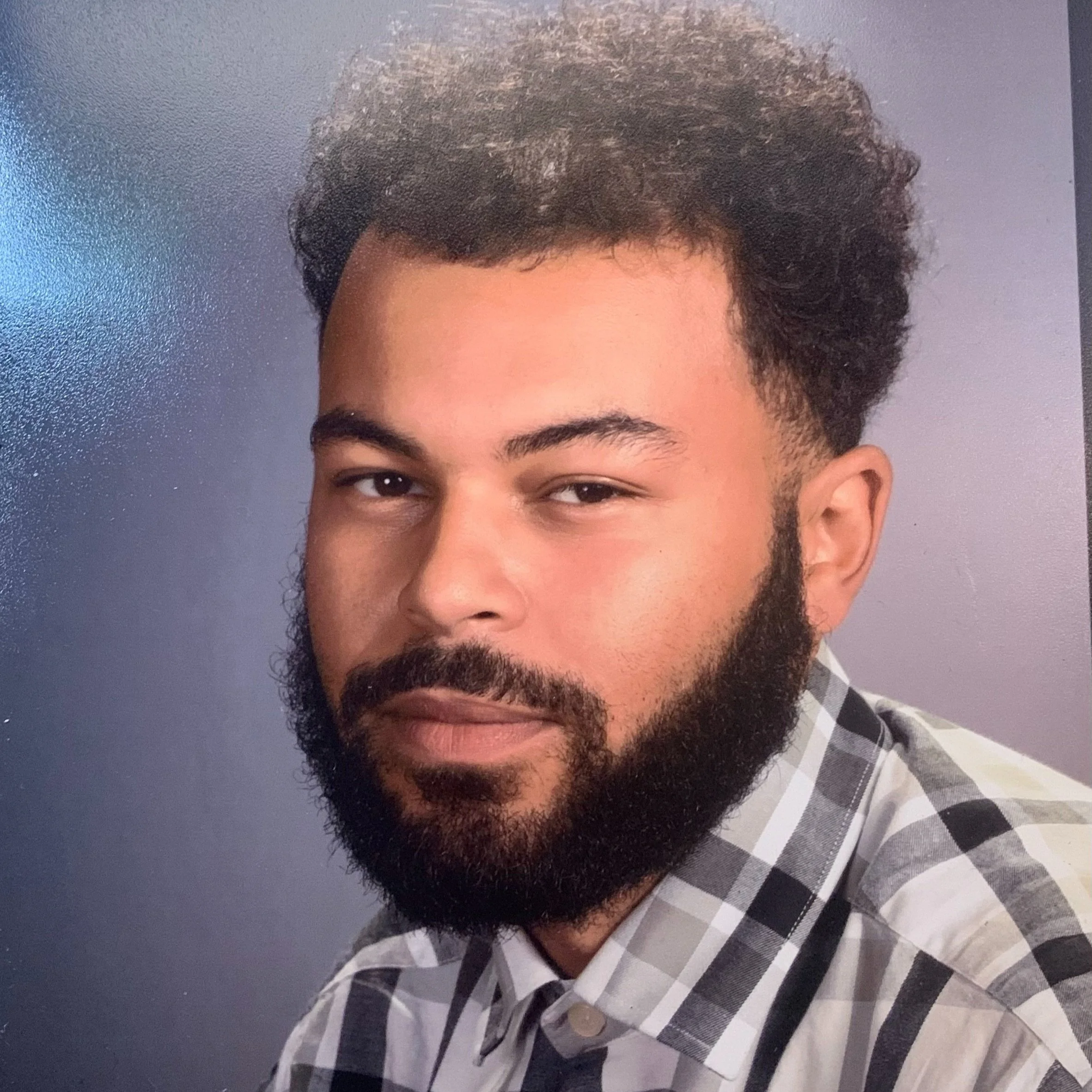 Portrait of a man with curly hair and a beard, wearing a checkered shirt and facing the camera with a neutral expression against a plain background.