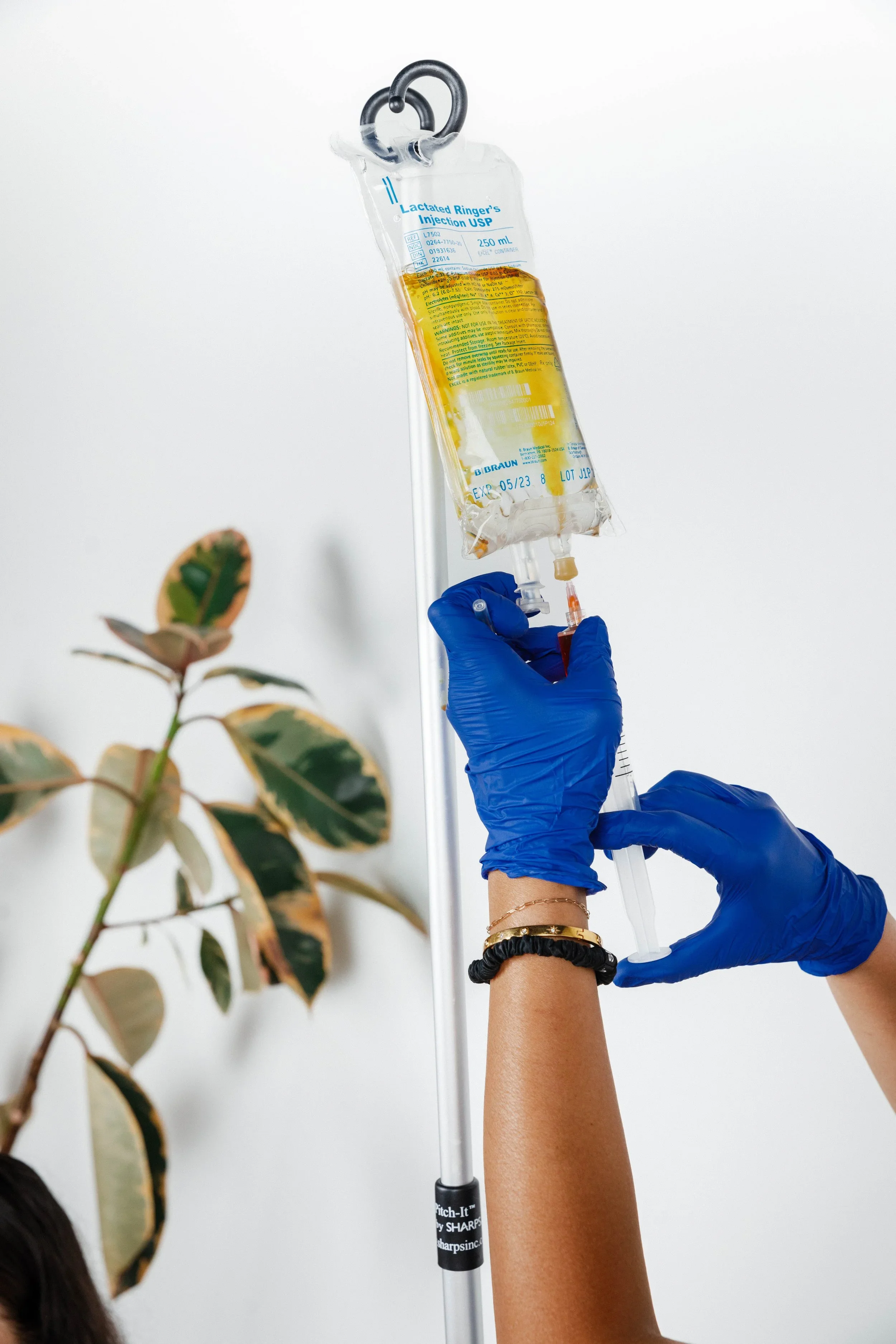 A person wearing blue gloves administering an injection from a syringe into a bag of intravenous fluid hanging from a stand.