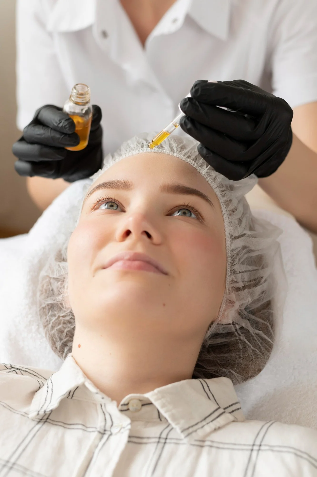 A woman receiving a cosmetic facial treatment with a syringe, lying down with a hair cap, while a practitioner in black gloves administers a liquid from a small bottle.