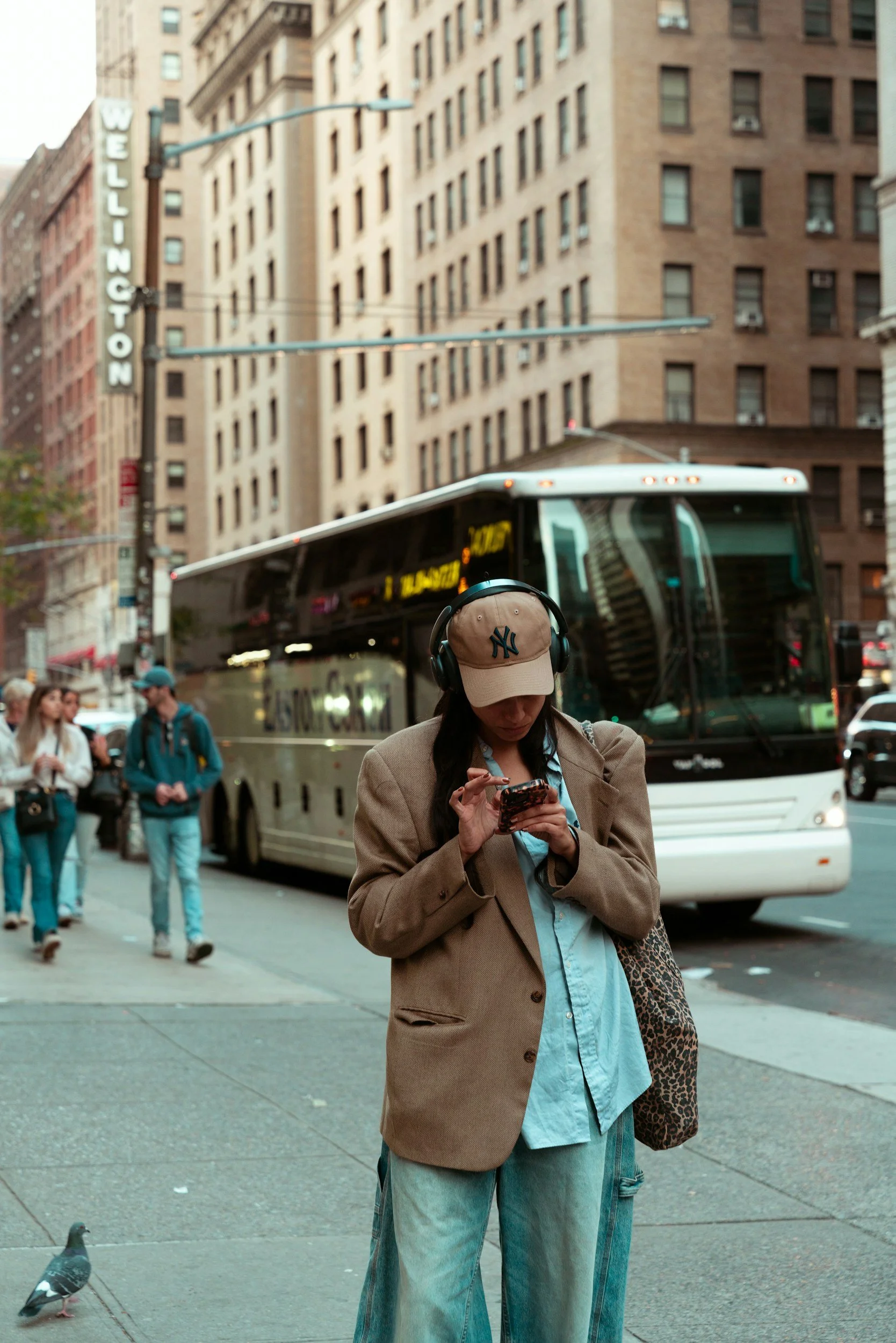 A woman wearing a beige baseball cap with a New York Yankees logo, a beige blazer, and light blue shirt, standing on a city sidewalk and looking at her phone. There is a busy urban street scene with a bus, several pedestrians, and tall buildings in the background.