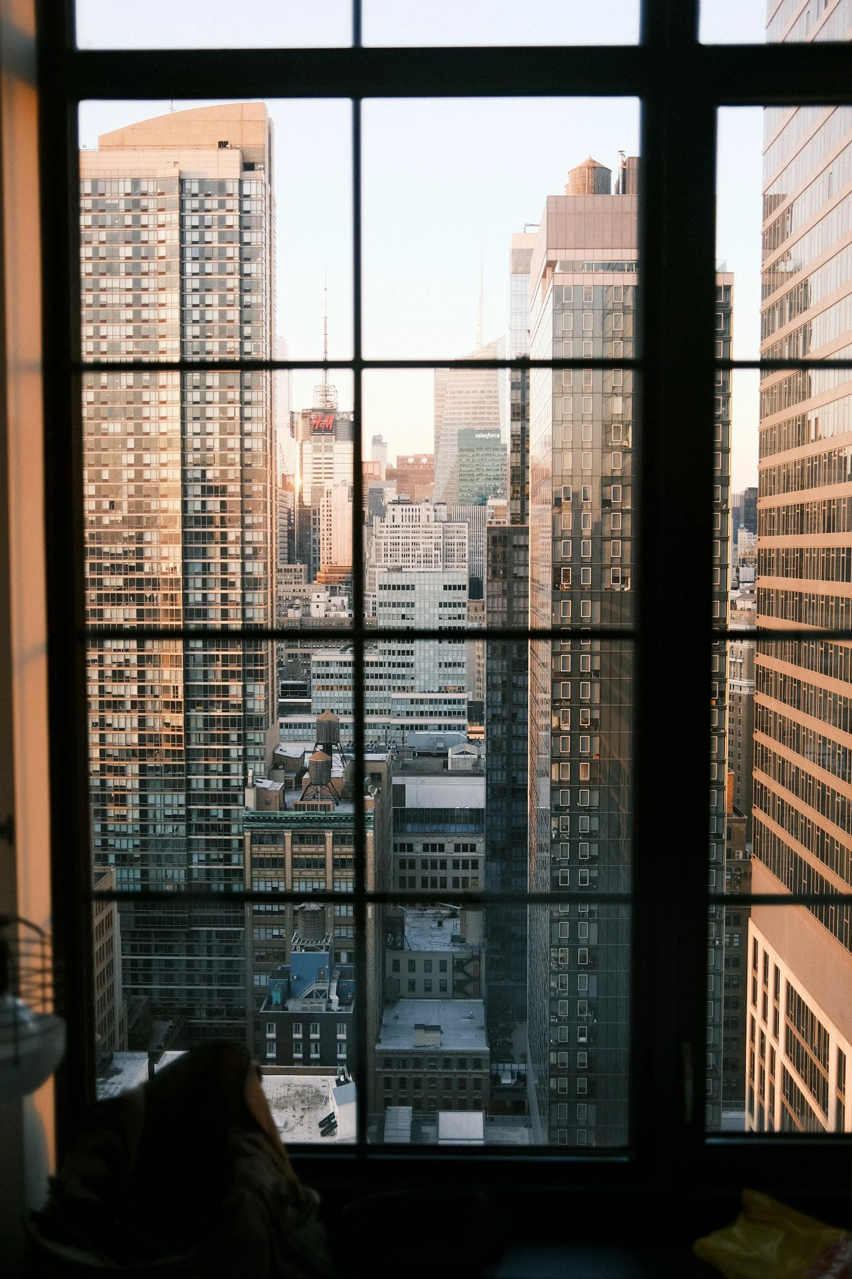Cityscape view through a window with black frames, showing tall modern skyscrapers in a dense urban area. The photo appears to be taken from inside a building, with a person visible at the bottom left corner.