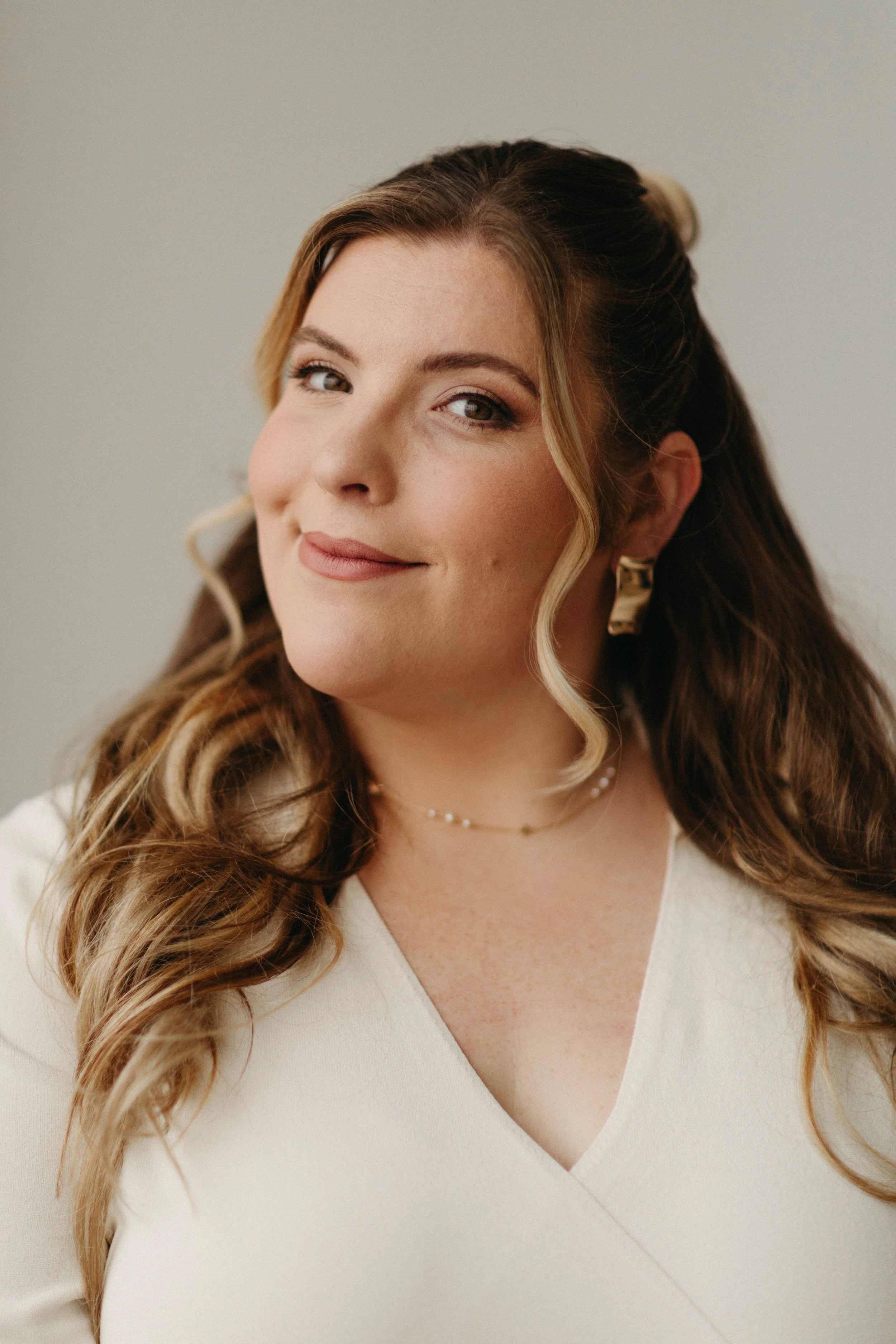 Close-up of a woman with long wavy hair, wearing a cream-colored V-neck top and gold jewelry, smiling softly against a plain background.