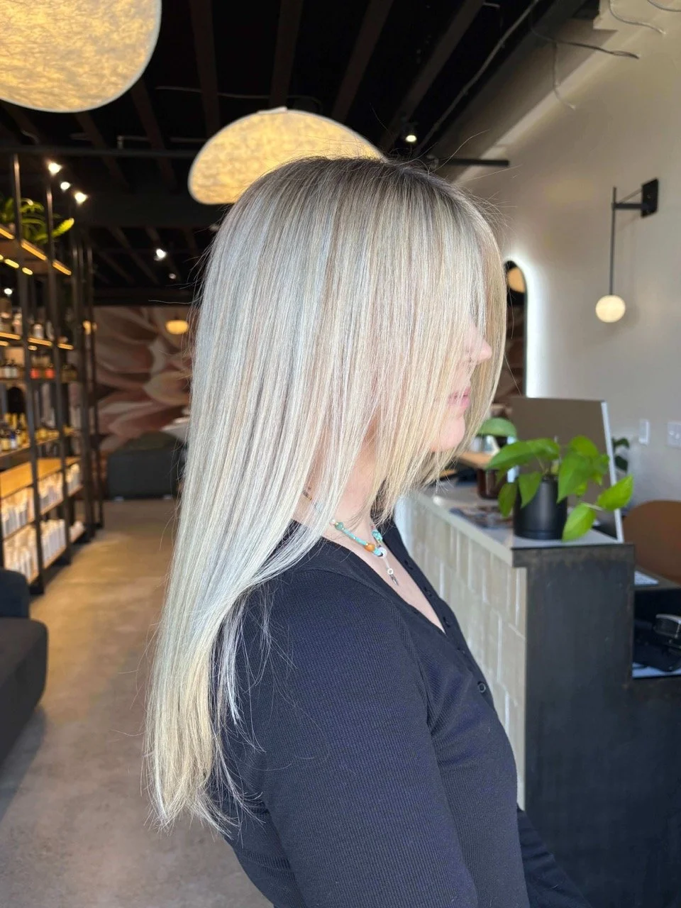 Woman with long, straight, platinum blonde hair indoors at a modern space with black walls and ceiling, pendant lights, and potted plants.