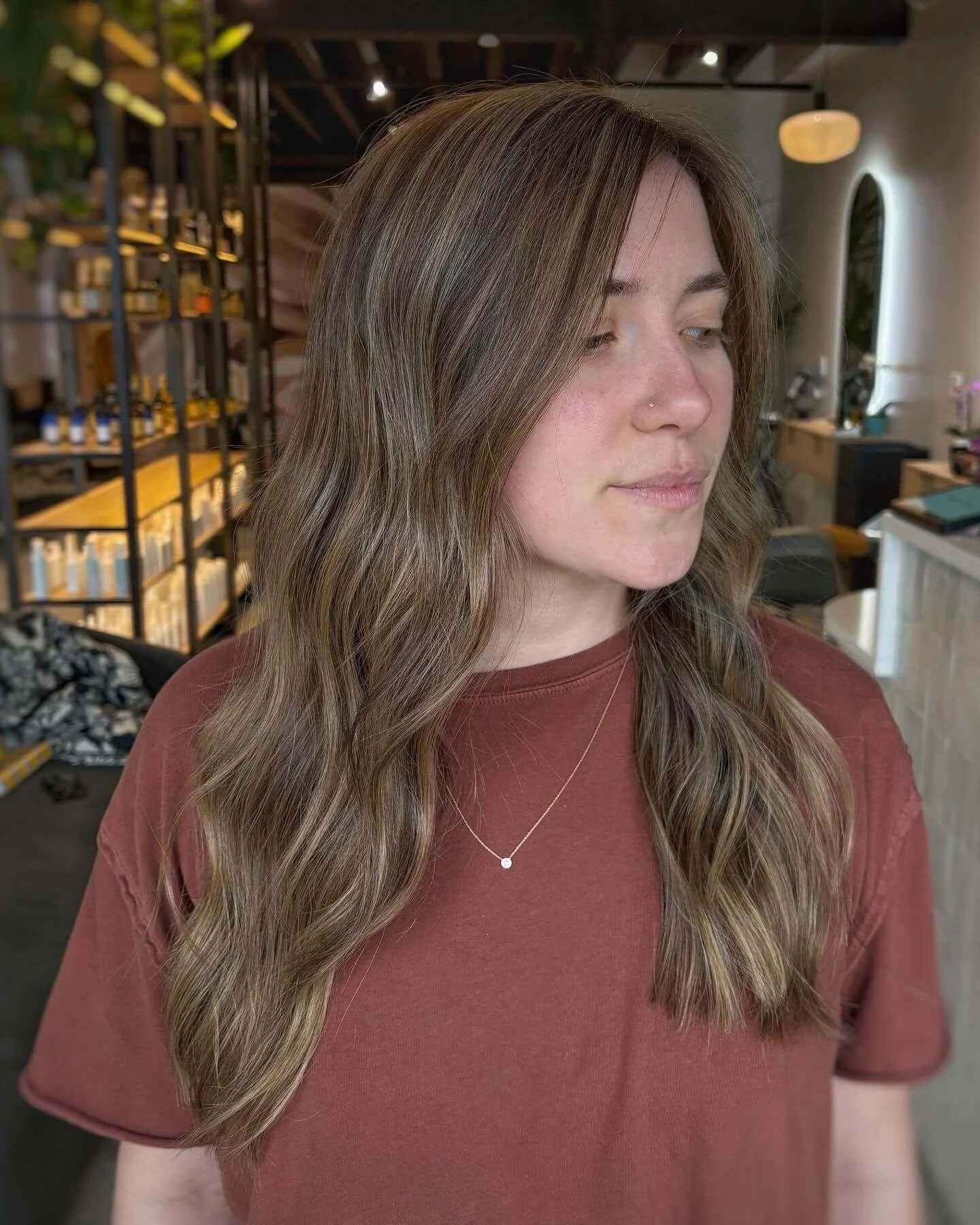 A woman with wavy, shoulder-length brown hair stands indoors, wearing a red t-shirt and a delicate necklace. The background features a modern interior with shelving, plants, and contemporary decor.
