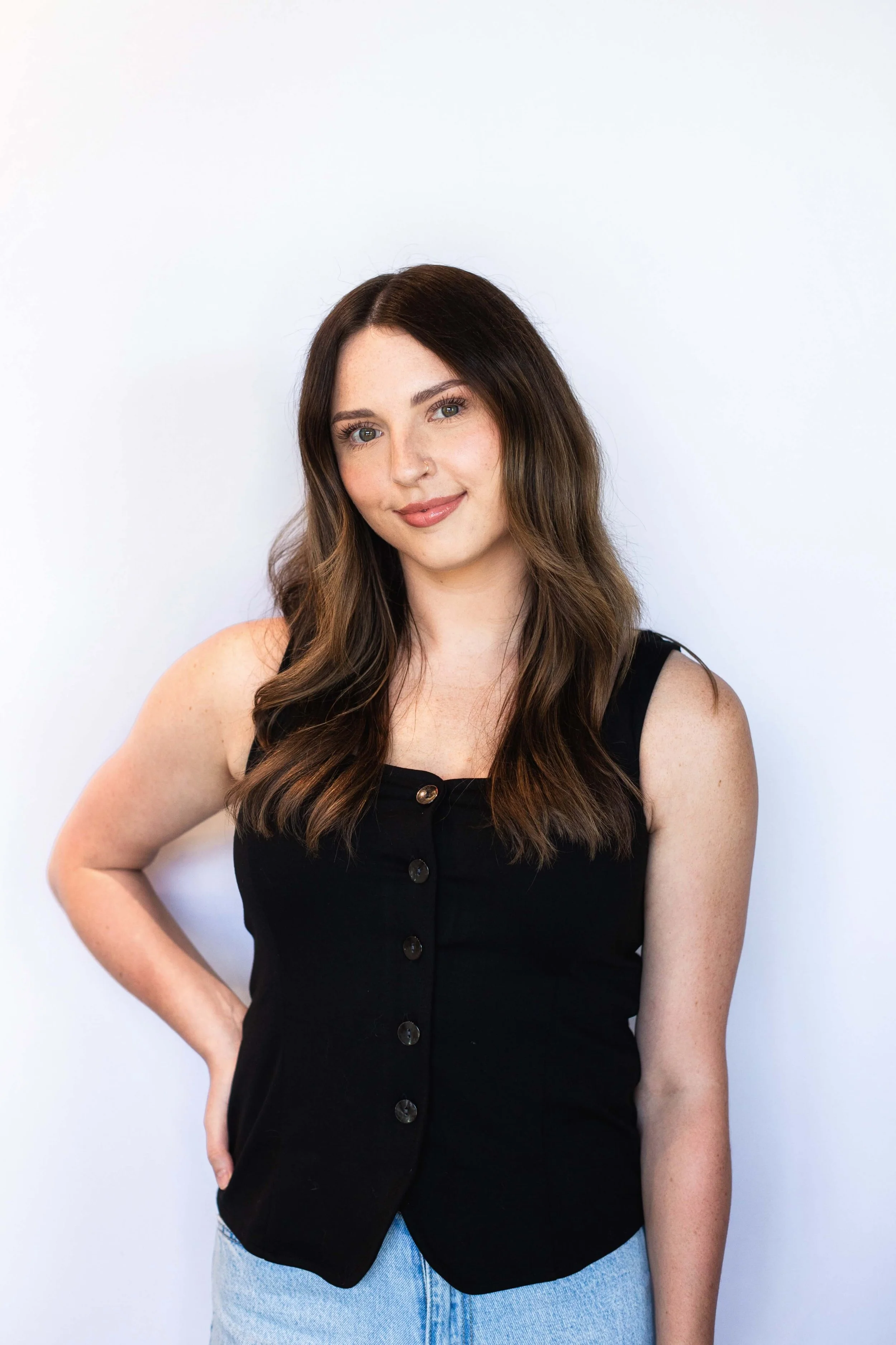 Taylor with long wavy brown hair, wearing a sleeveless black button-up top and light blue jeans, standing against a plain white background.