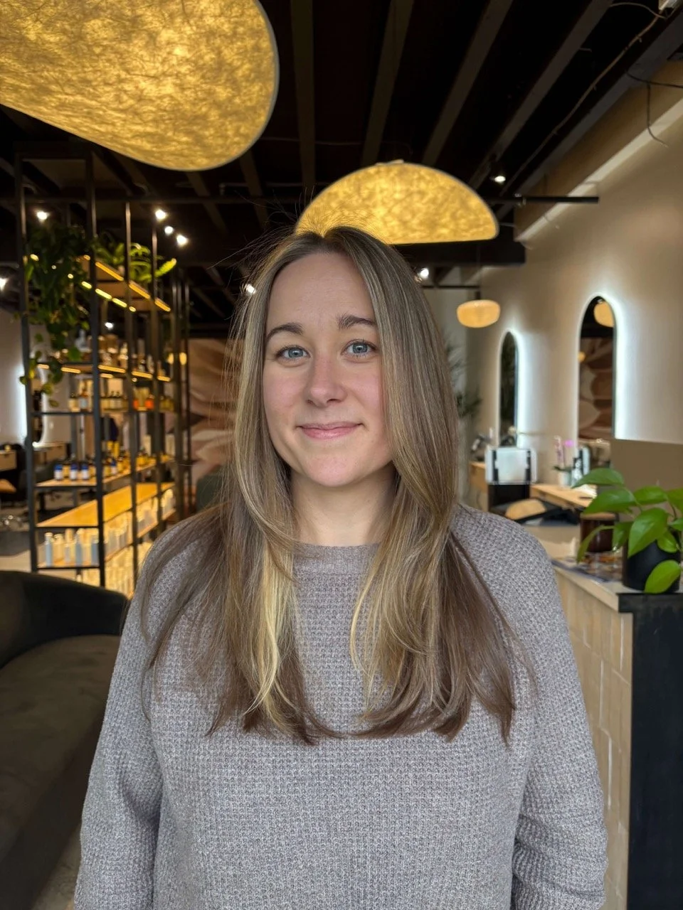A woman with long, light brown hair and blue eyes smiling in a modern, well-lit indoor space with decorative yellow lighting fixtures and plants.