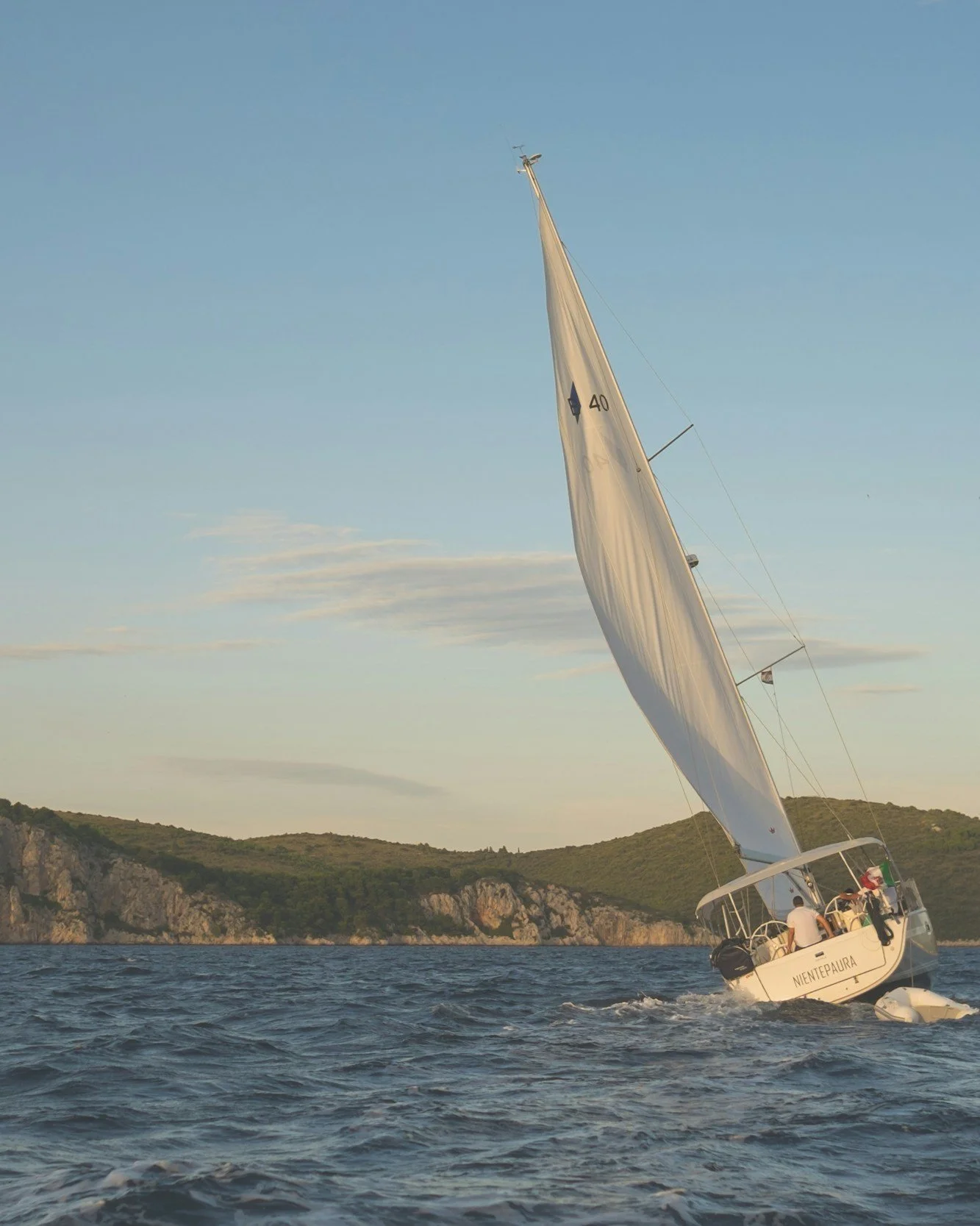 A sailboat tilted to the right with people on board, sailing on the water near a coastal landscape with hills and cliffs at sunset.
