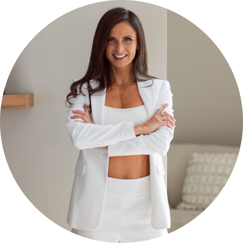 A woman in a white blazer and matching pants with a white crop top, smiling and crossing her arms, standing in a modern room with neutral walls and a patterned cushion in the background.