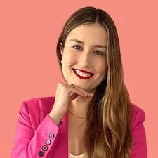 A woman with long brown hair and red lipstick smiling, wearing a pink blazer against a pink background.