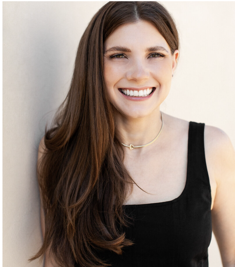 A woman with long brown hair and a bright smile, wearing a black sleeveless top and a necklace, standing against a light-colored wall.