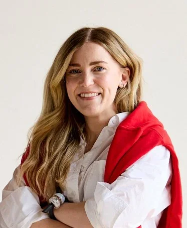 A woman with long blonde hair, wearing a white shirt and a red jacket draped over her shoulders, smiling with her arms crossed.