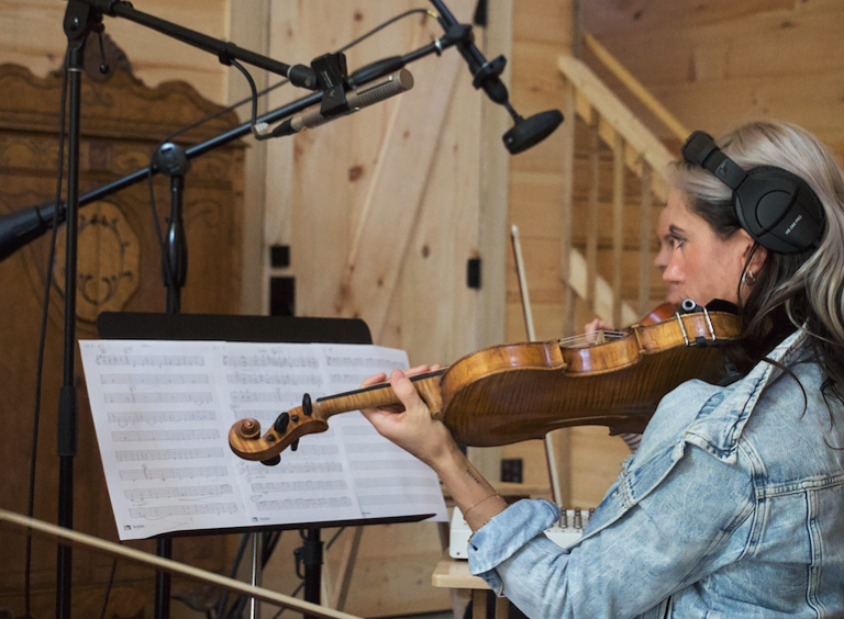 A woman playing the violin wearing large headphones in a recording studio with wooden walls.