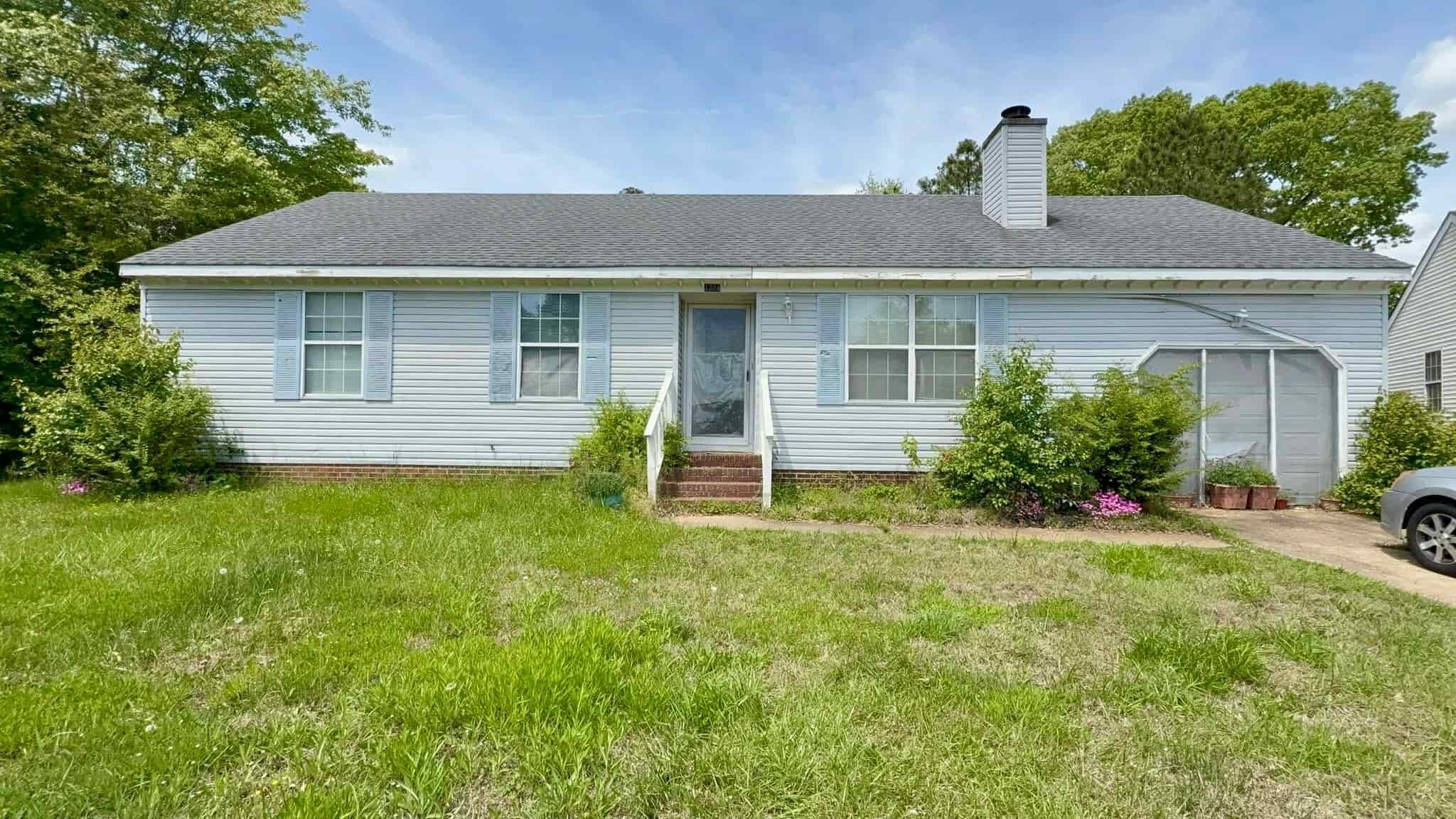Single-story white house with blue shutters, a porch with stairs, a garage on the right, and a lawn with bushes and trees in the background.