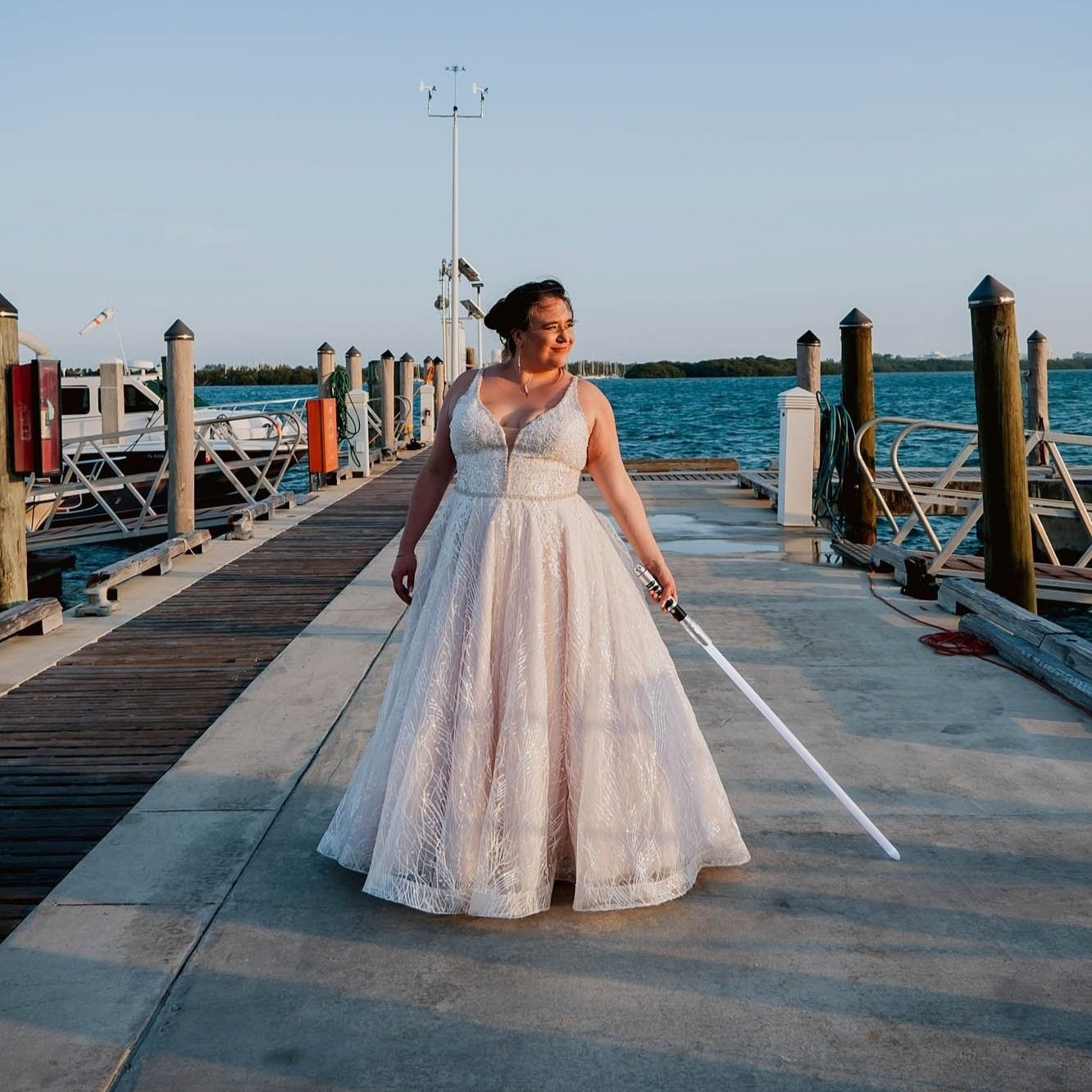 wedding portrait of bride standing on a pier holding a lightsaber during golden hour taken by Sharma Shari Photography