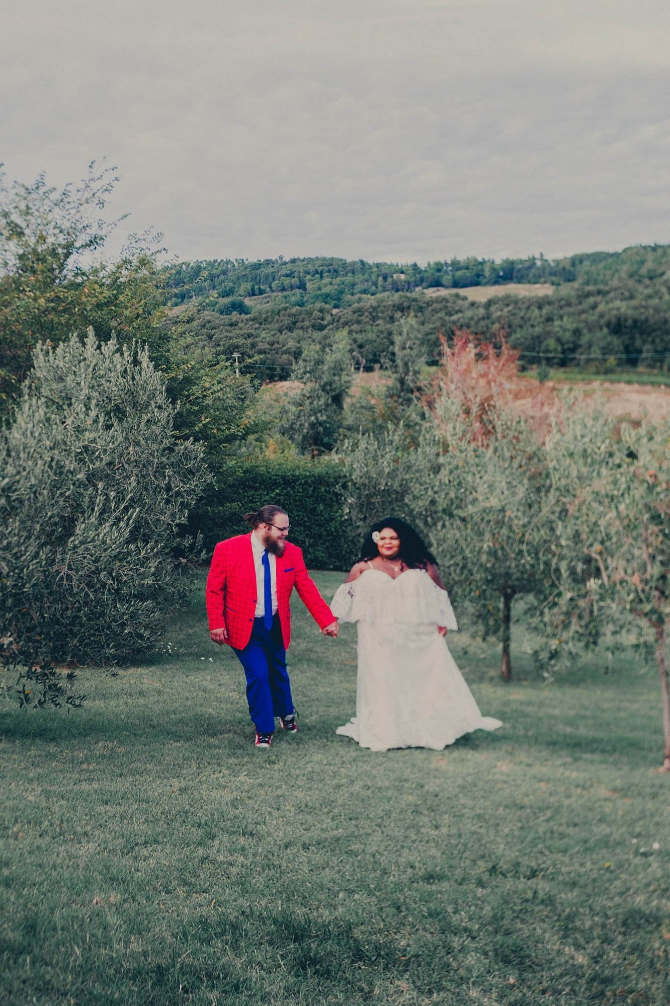 newlyweds in white gown and red blazer hold hands in a vineyard taken by Sharma Shari Photography