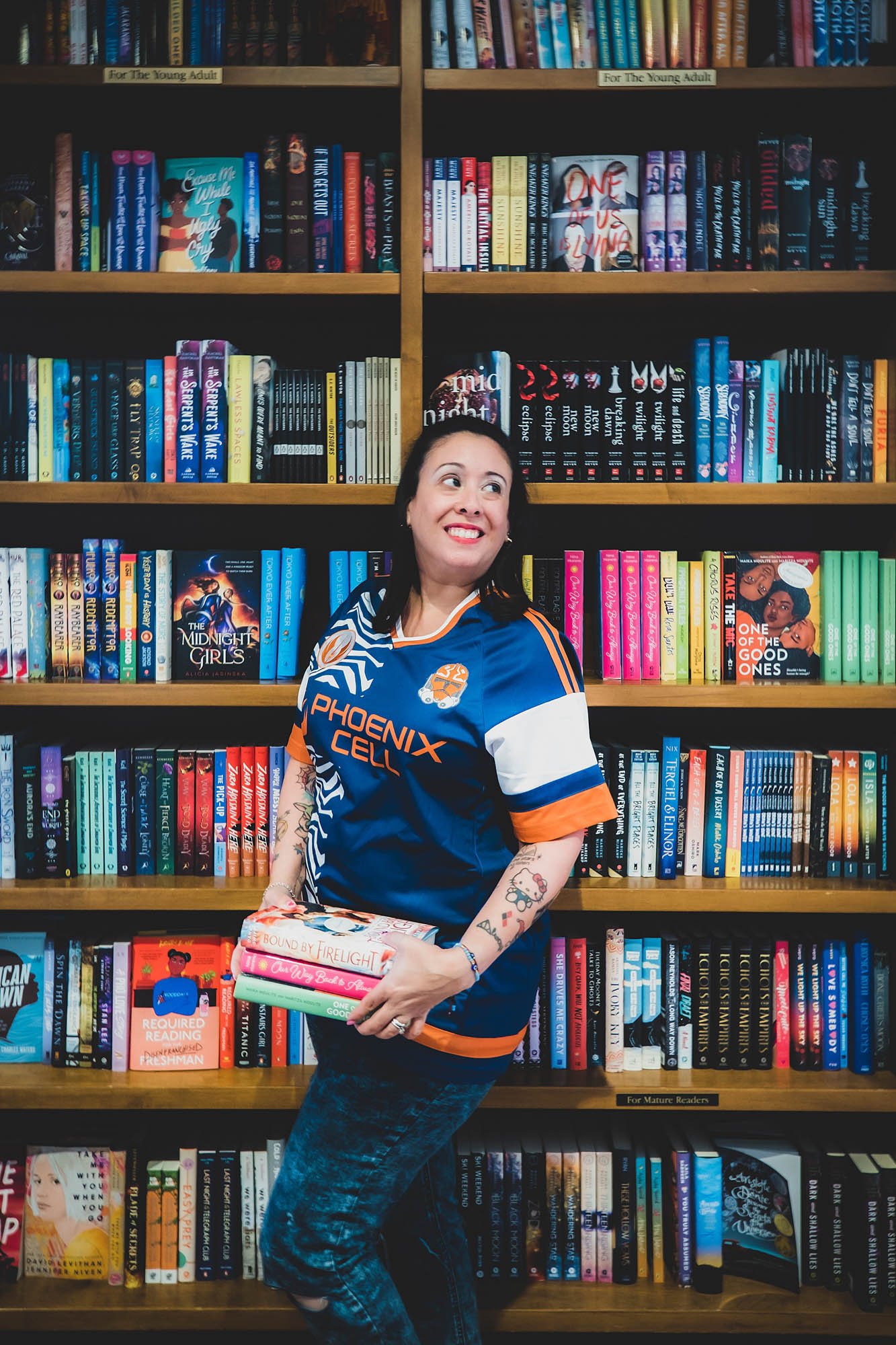 small business owner poses in front of bookshelves holding 3 books and smiling taken by Sharma Shari Photography