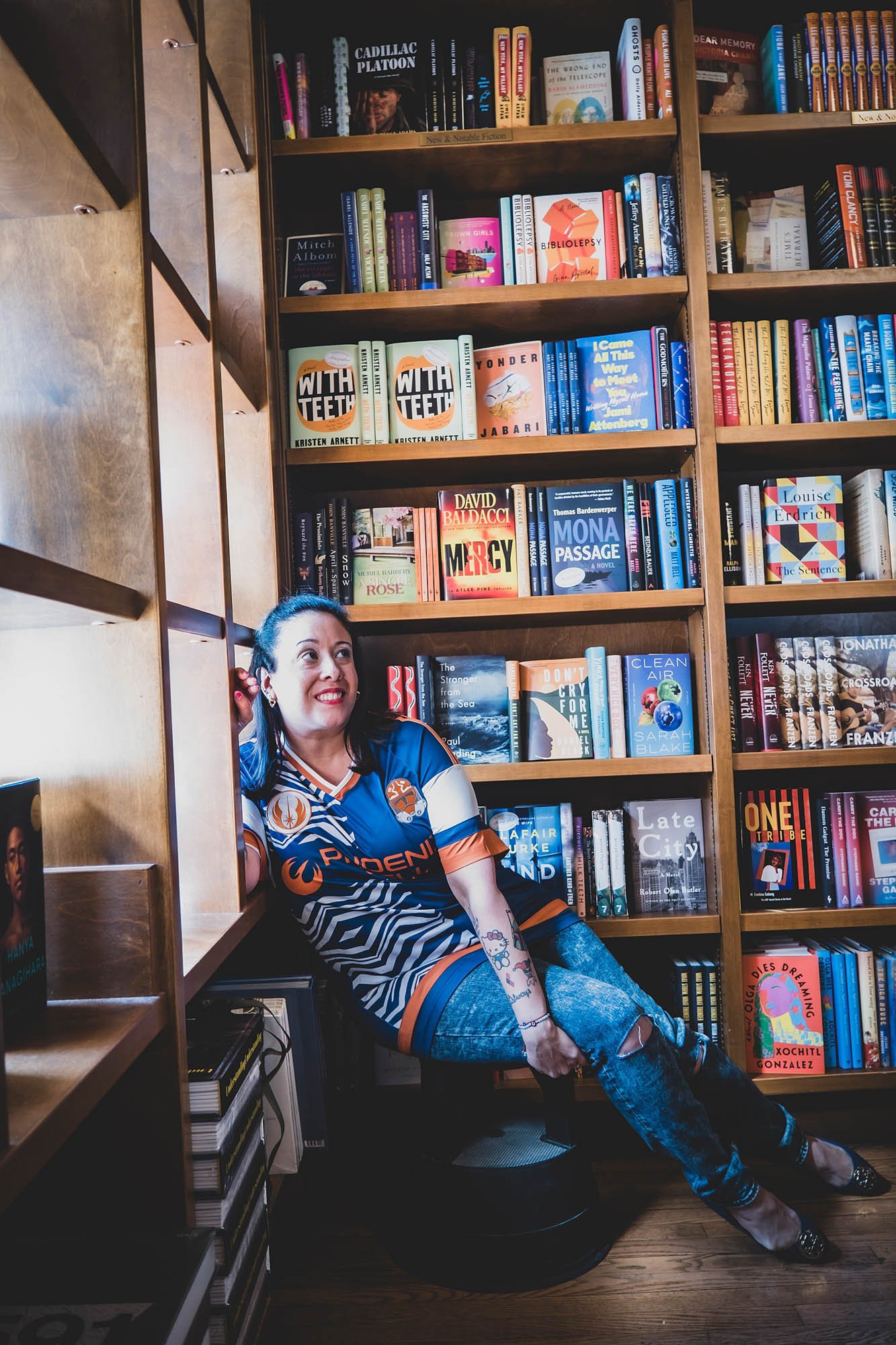 shop owner smiles in front of and leans on full book shelves taken by Sharma Shari Photography