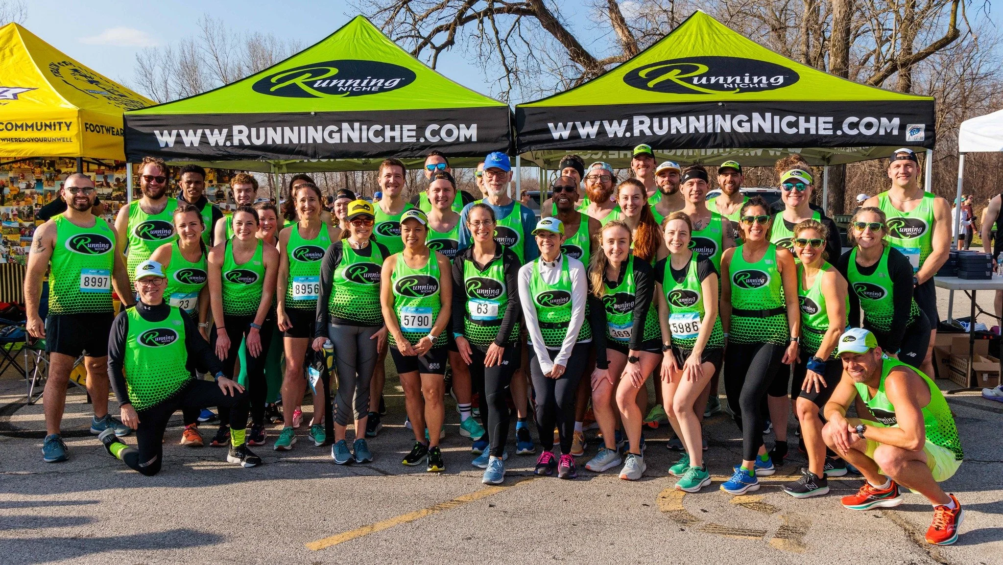 Group of runners wearing green and black uniforms, with race numbers, gathered in front of ‘Running Niche’ branded tents during a community running event.