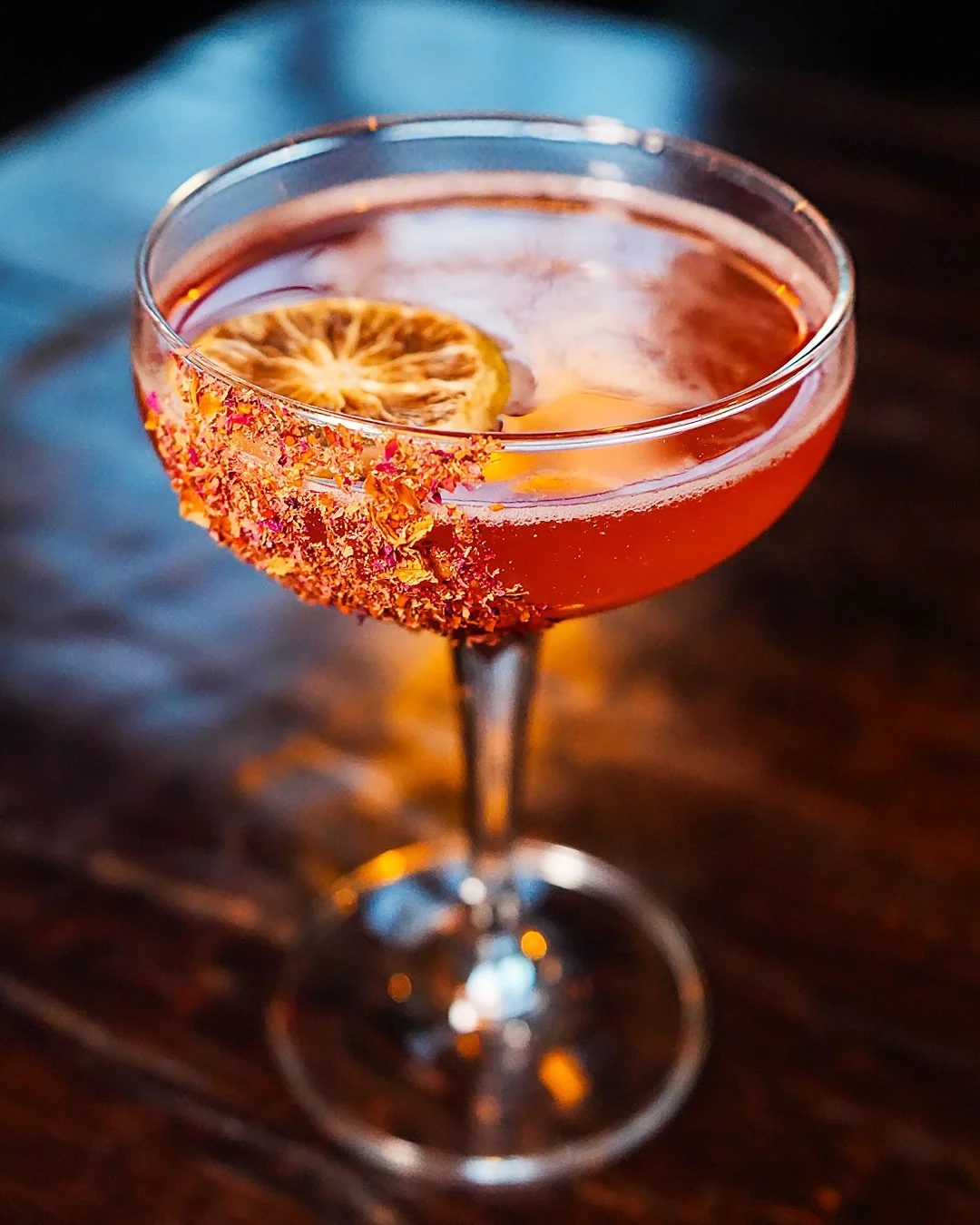 A cocktail in a coupe glass with a salted rim and dried lemon slice garnish, on a dark wooden surface.