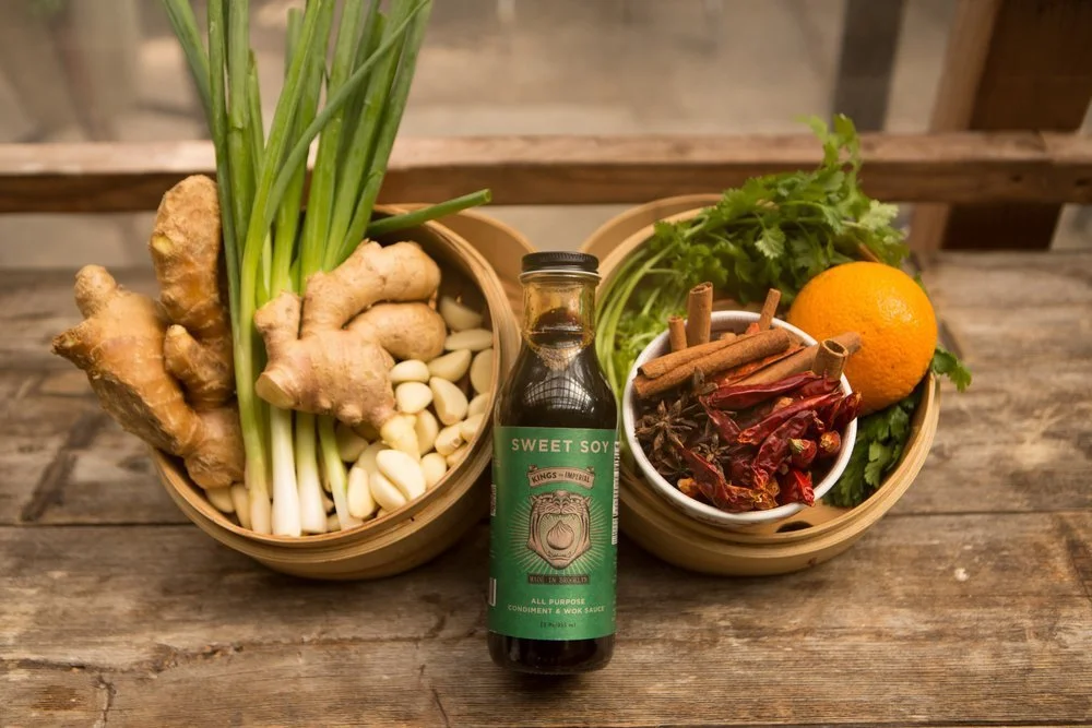 Baskets with fresh ginger, garlic, green onions, cilantro, dried red chili peppers, cinnamon sticks, an orange, and a bottle of sweet soy sauce on a wooden surface.