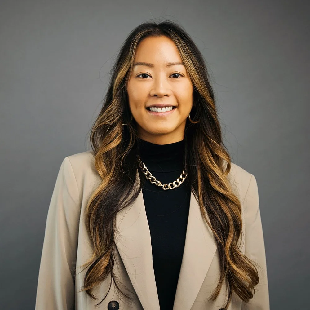 A woman with long wavy hair wearing a beige blazer, black top, gold chain necklace, and gold hoop earrings, smiling against a gray background.