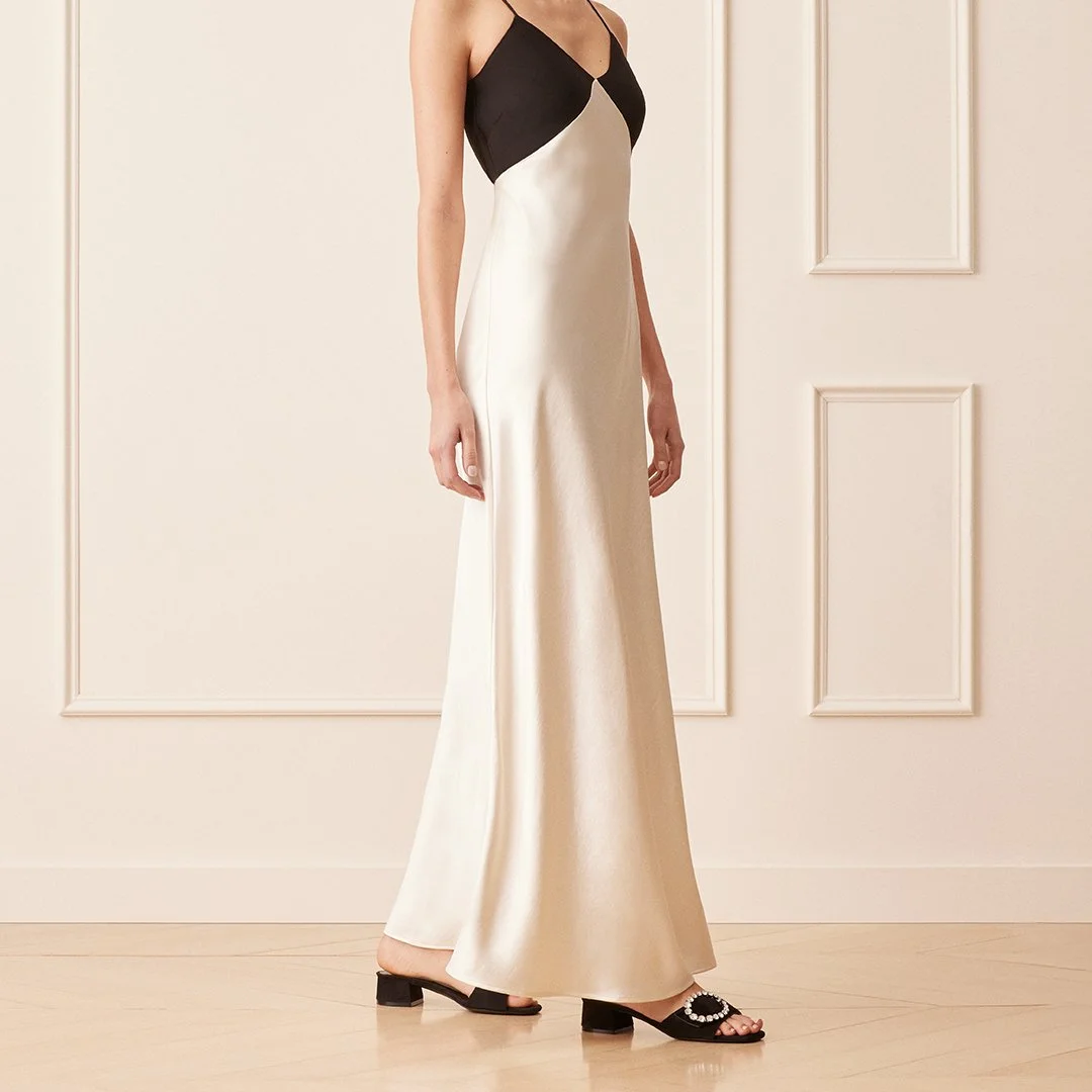 A woman wearing a sleeveless, color-block dress with black on top and cream on the bottom, paired with black heeled sandals with decorative embellishments, standing in a room with cream walls and wooden floors.