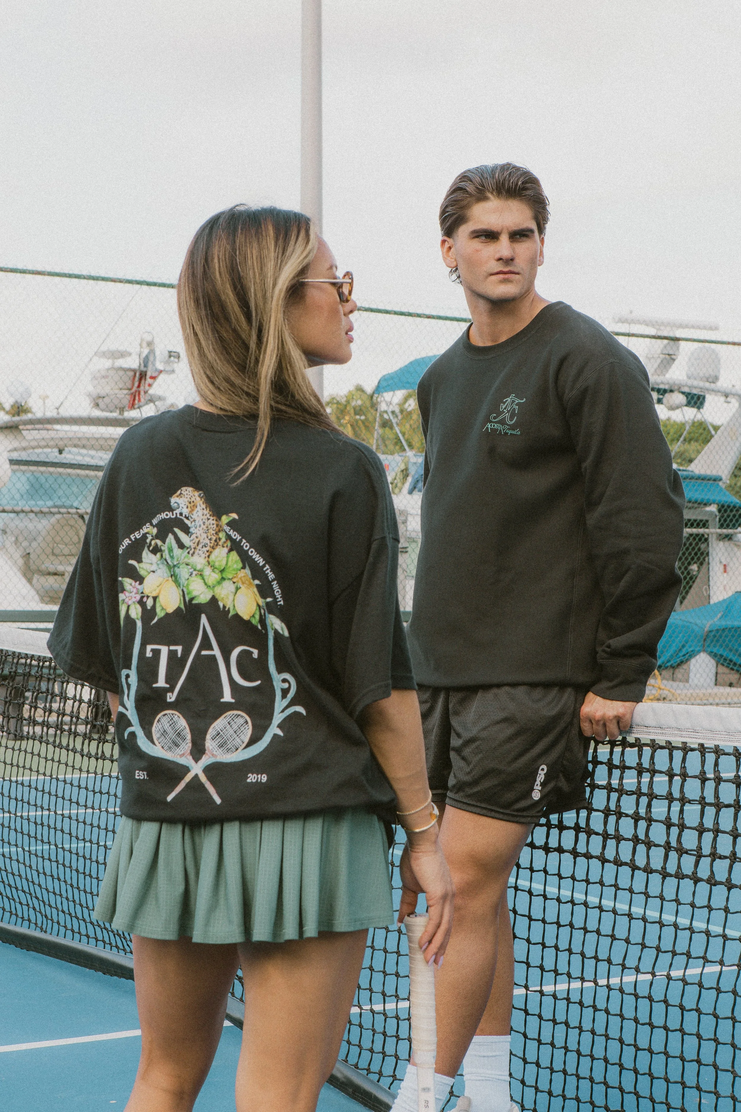 Two young people, a woman and a man, standing on a tennis court with a net in the foreground. The woman is wearing glasses, a black shirt with a graphic on the back, and a green skirt. The man is wearing a black sweatshirt and black shorts, holding a tennis racket, with airplanes in the background.