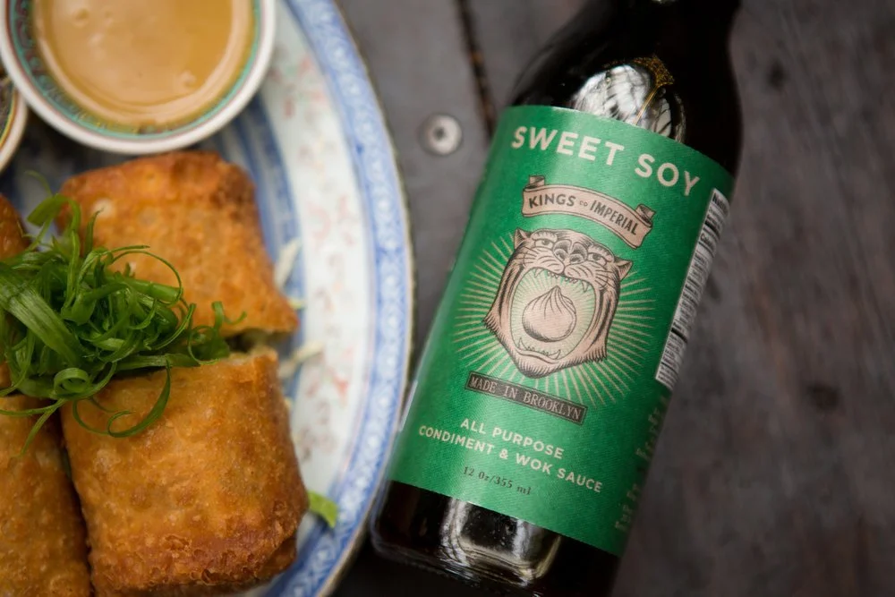 Bottle of Sweet Soy Kings Imperial all-purpose condiment and wok sauce next to a plate of spring rolls garnished with green onions, on a dark wooden surface.