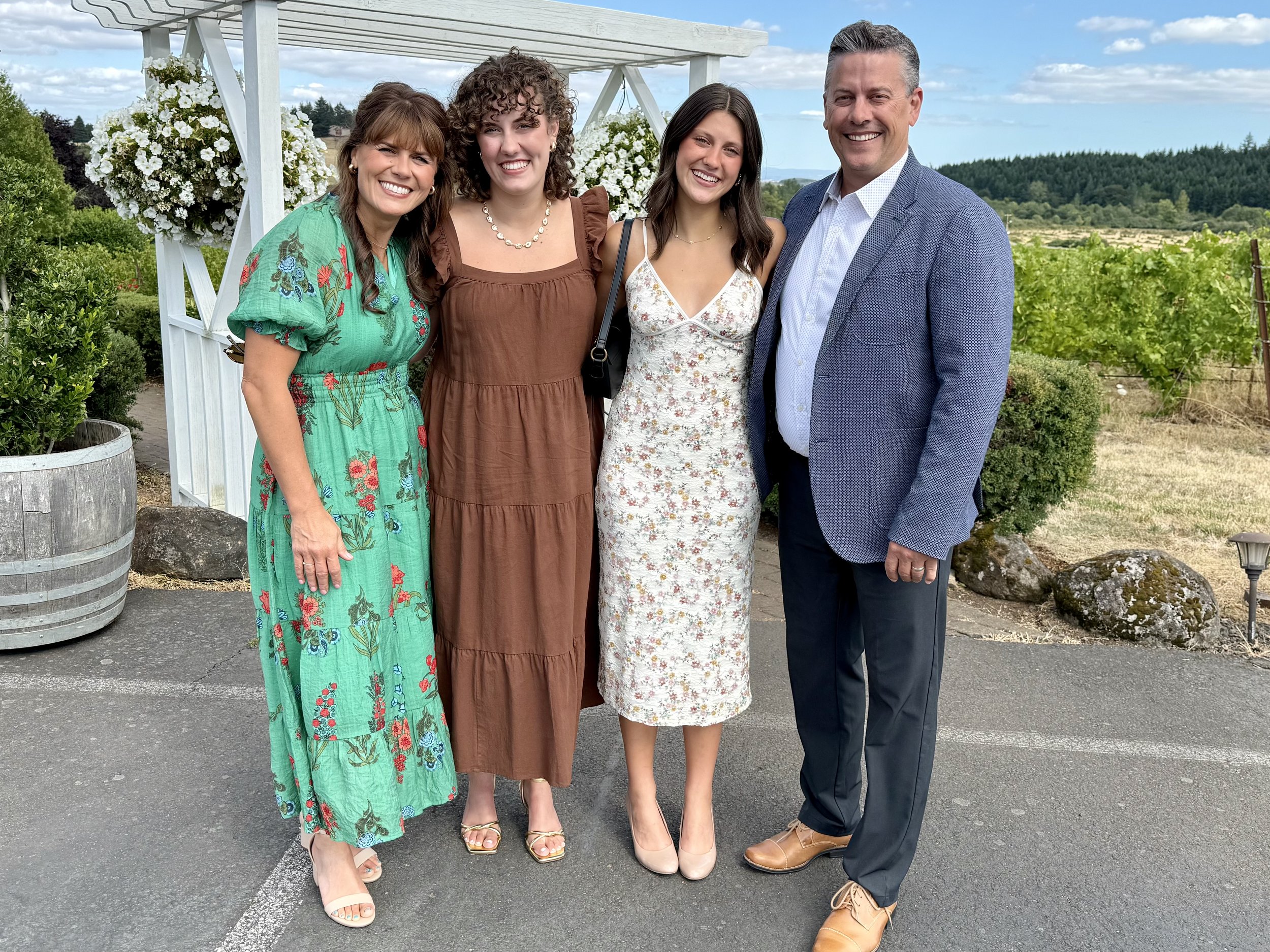 Four women and one man standing outside near white wooden arch decorated with white flowers, with green bushes, trees, and rural landscape in the background. They are smiling and dressed in semi-formal summer clothes.