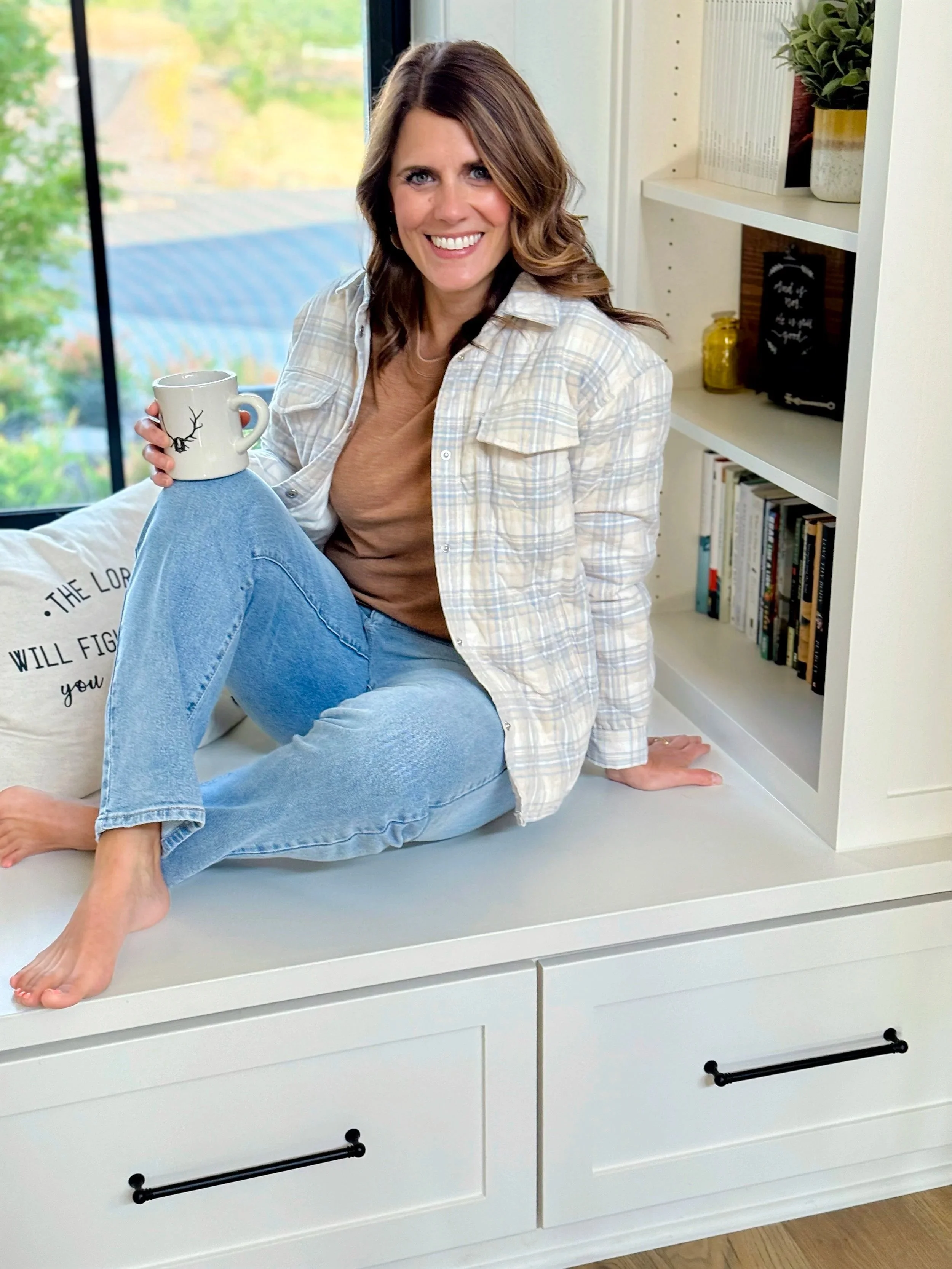A smiling woman with wavy brown hair sitting on a white cabinet near a window, holding a white mug with a black reindeer design. She is wearing a beige t-shirt and a plaid button-up shirt, with a bookshelf and a potted plant visible in the background.