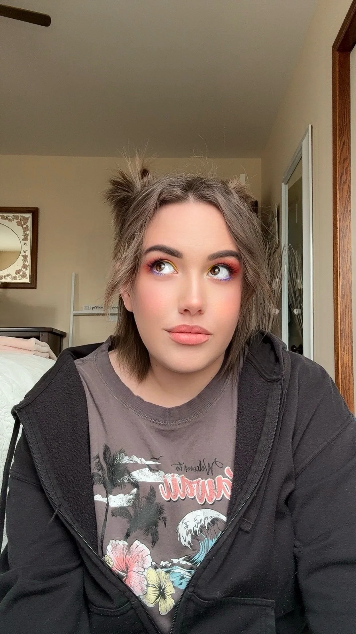 A young woman with short, tousled hair and colorful eye makeup, wearing a graphic t-shirt and black jacket, sitting indoors with a neutral expression.